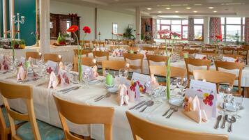 A large dining hall with multiple tables set for a formal event, featuring wooden chairs and white tablecloths.