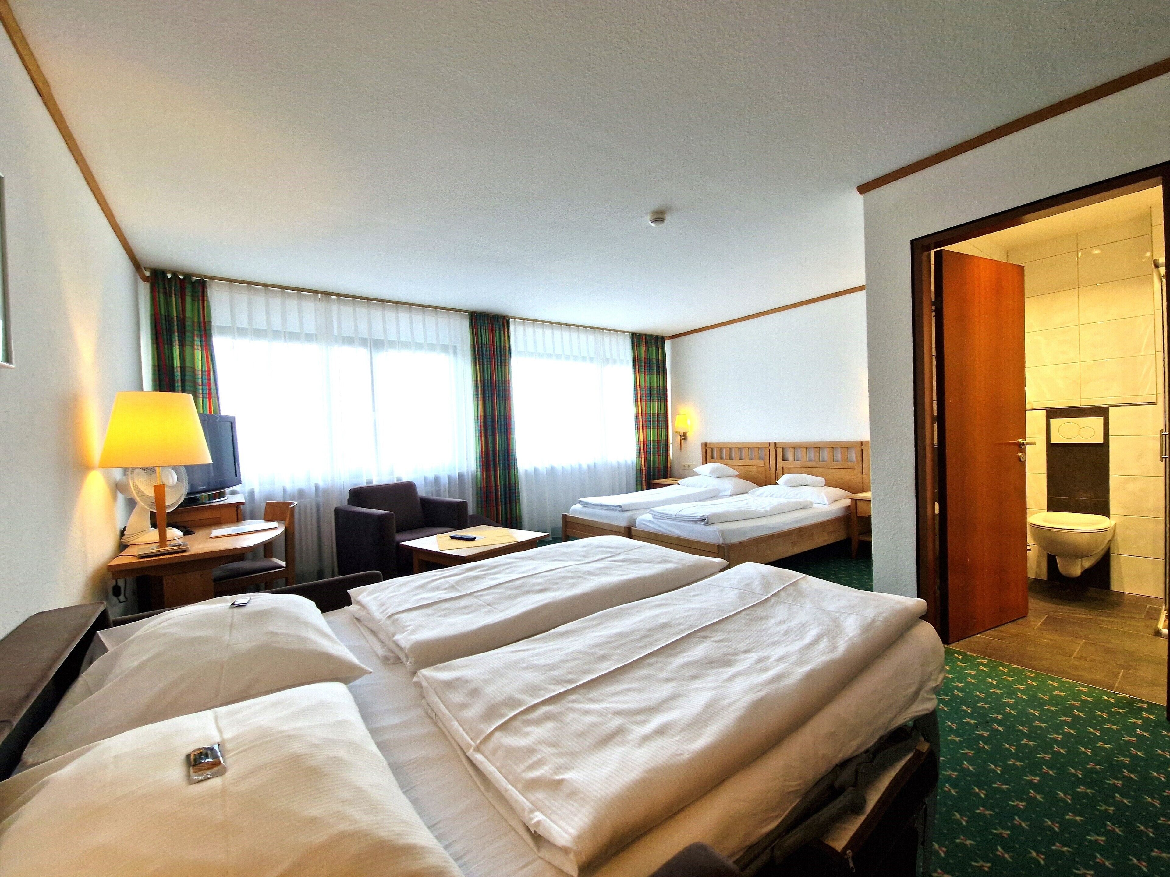 A hotel room with three beds, a desk, a chair, a TV, and a bathroom visible through an open door.