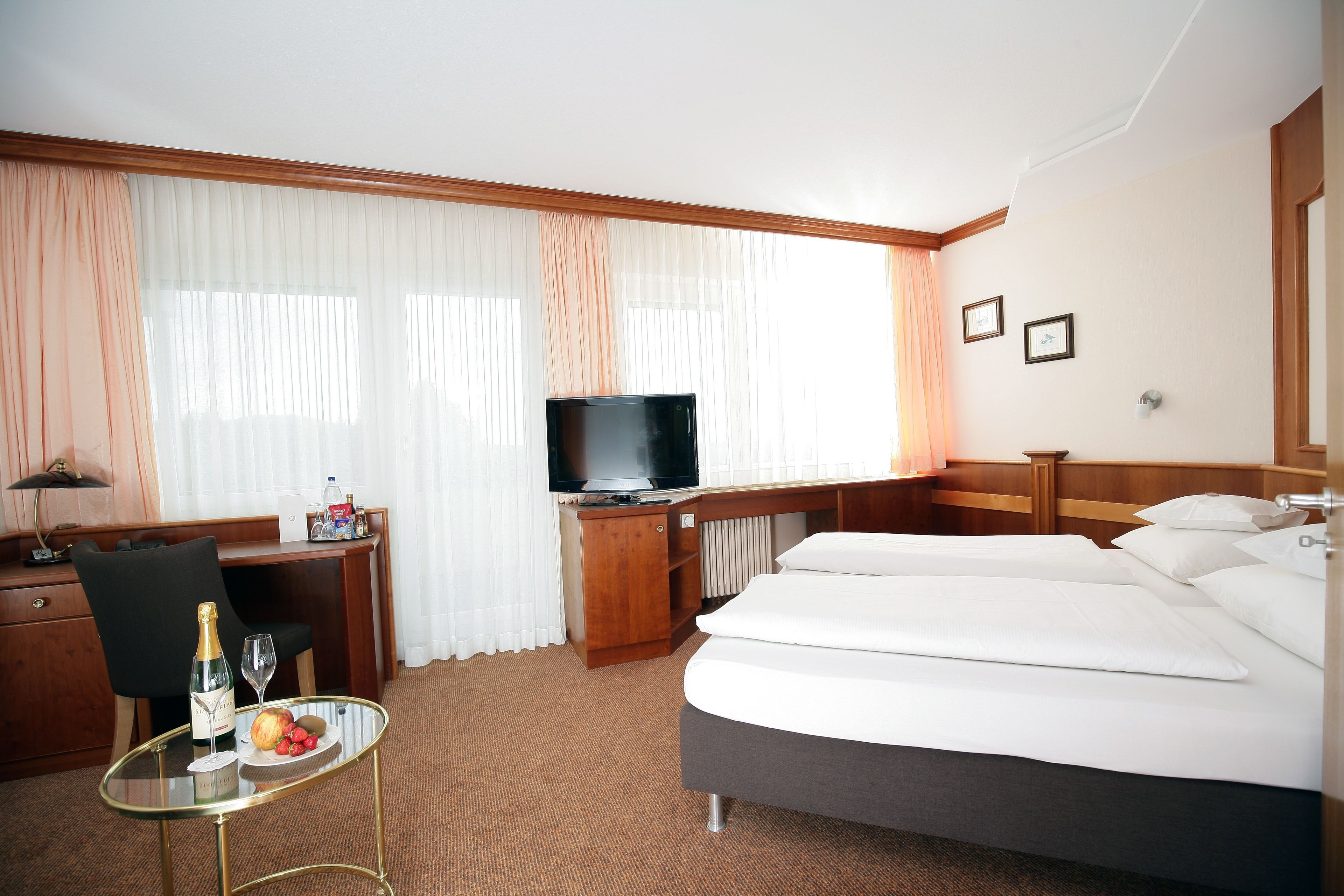 Ein Hotelzimmer mit einem Bett, einem Schreibtisch mit Fernseher, einem kleinen Tisch mit Flasche und Gläsern sowie einem Fenster mit Vorhängen.