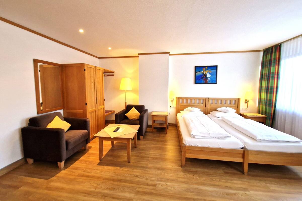 Habitación de hotel con cama, dos sillones, un armario de madera, una mesita pequeña y dos lámparas de noche.