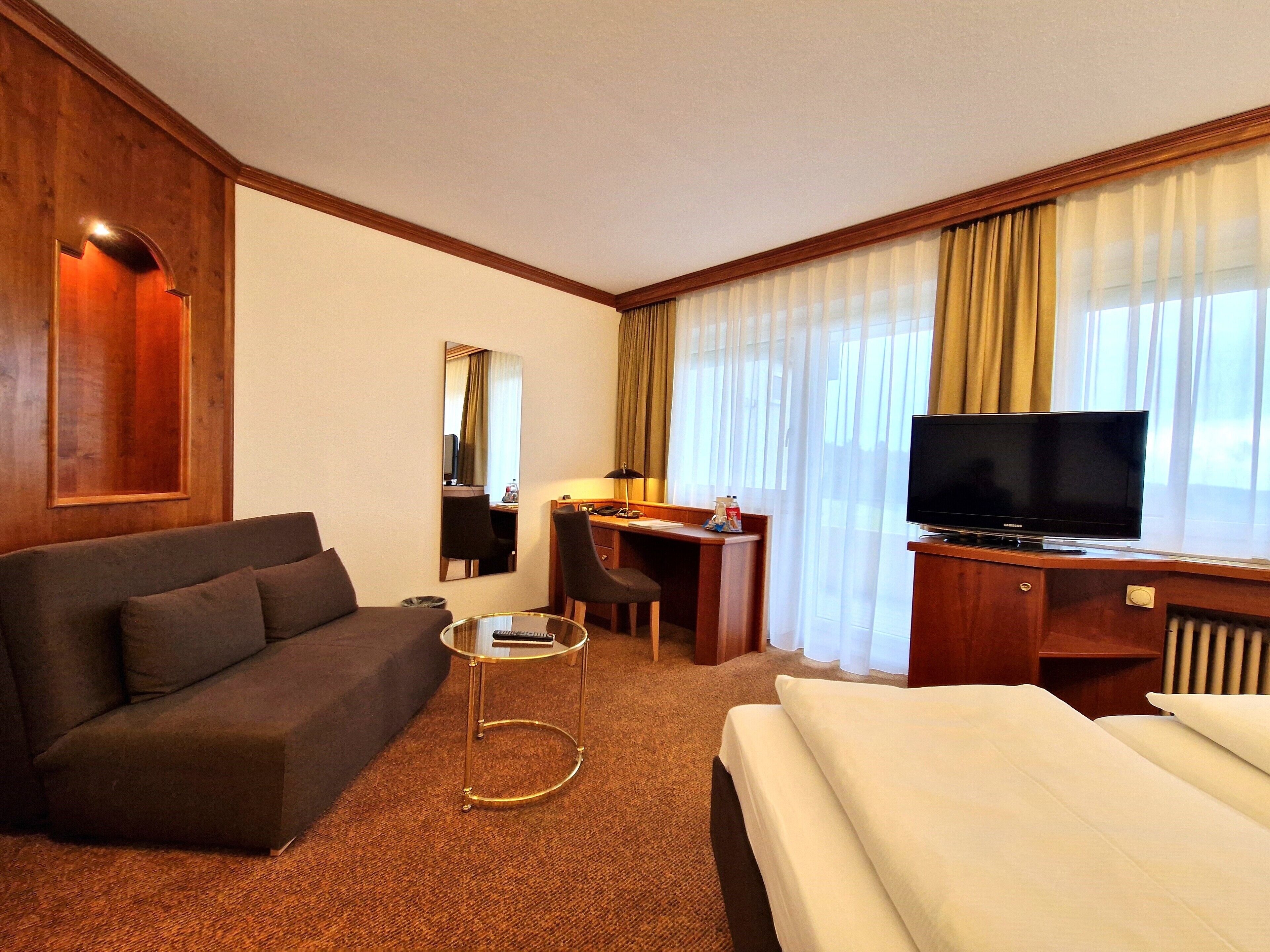 Habitación de hotel con cama, sofá, escritorio con silla, televisión y ventana con cortinas.