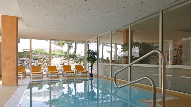 An indoor swimming pool with clear glass walls, a metal pool ladder, and several lounge chairs. - Badhotel Restaurant Stauferland (Bad Boll)