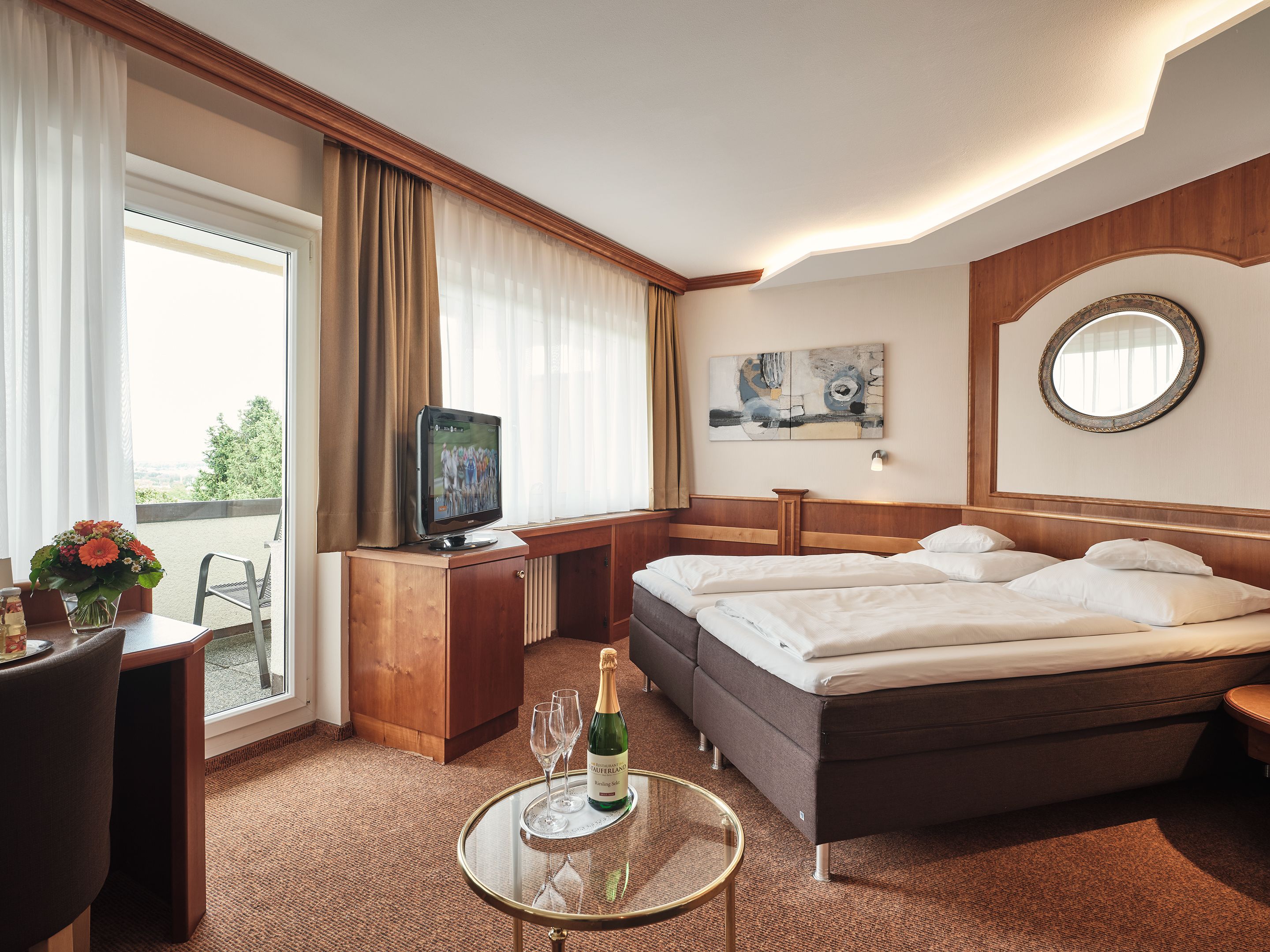 Ein Hotelzimmer mit Bett, Schreibtisch mit Fernseher, Tisch mit Champagnerflasche und Gläsern sowie Blick ins Freie.