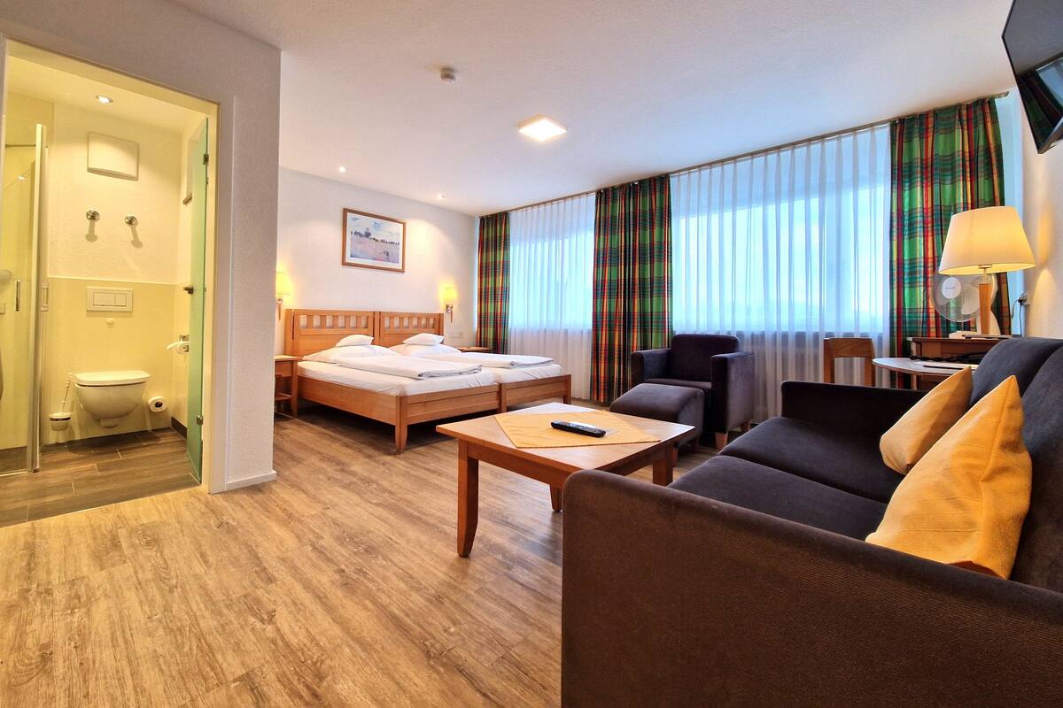 Habitación de hotel con piso de madera, una cama con ropa blanca, un sofá, una mesa y un televisor.