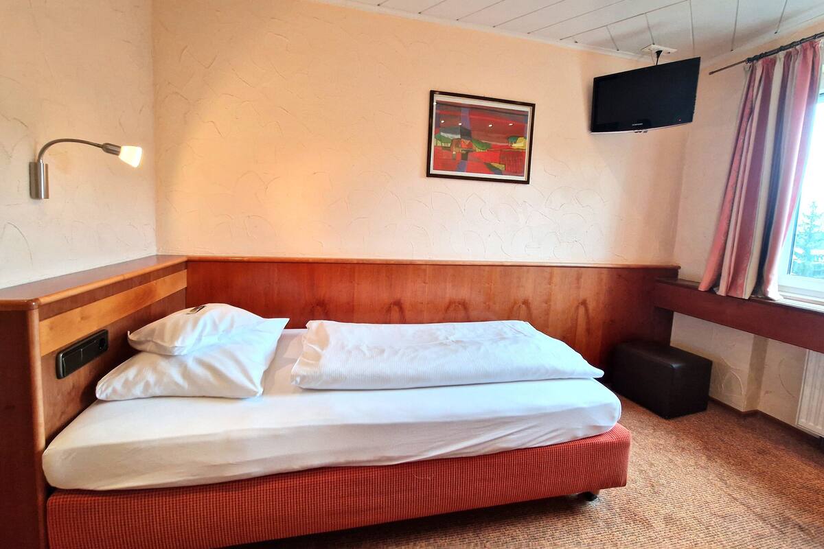 Una cama bien hecha con sábanas blancas, cabecera de madera, televisor de pared y ventana con cortinas.