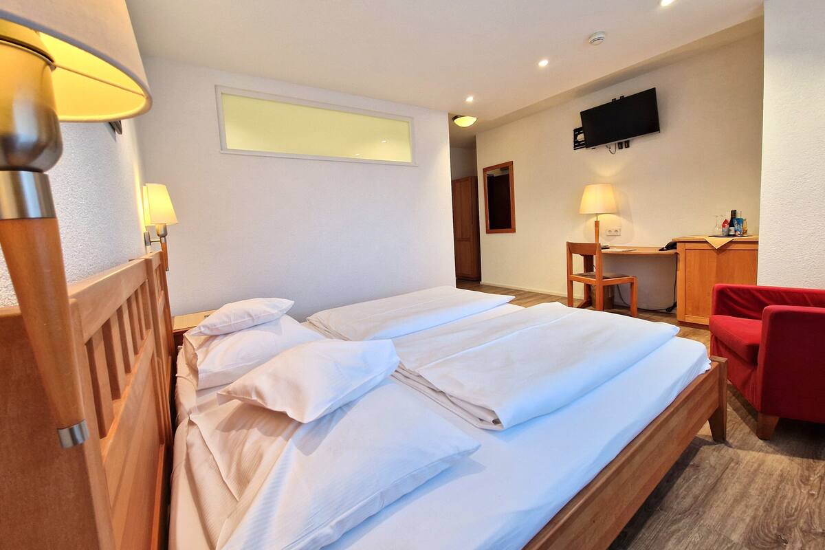 Una cama bien hecha con ropa blanca, un cabecero de madera y un escritorio con una silla en una habitación con paredes blancas y una lámpara de techo.