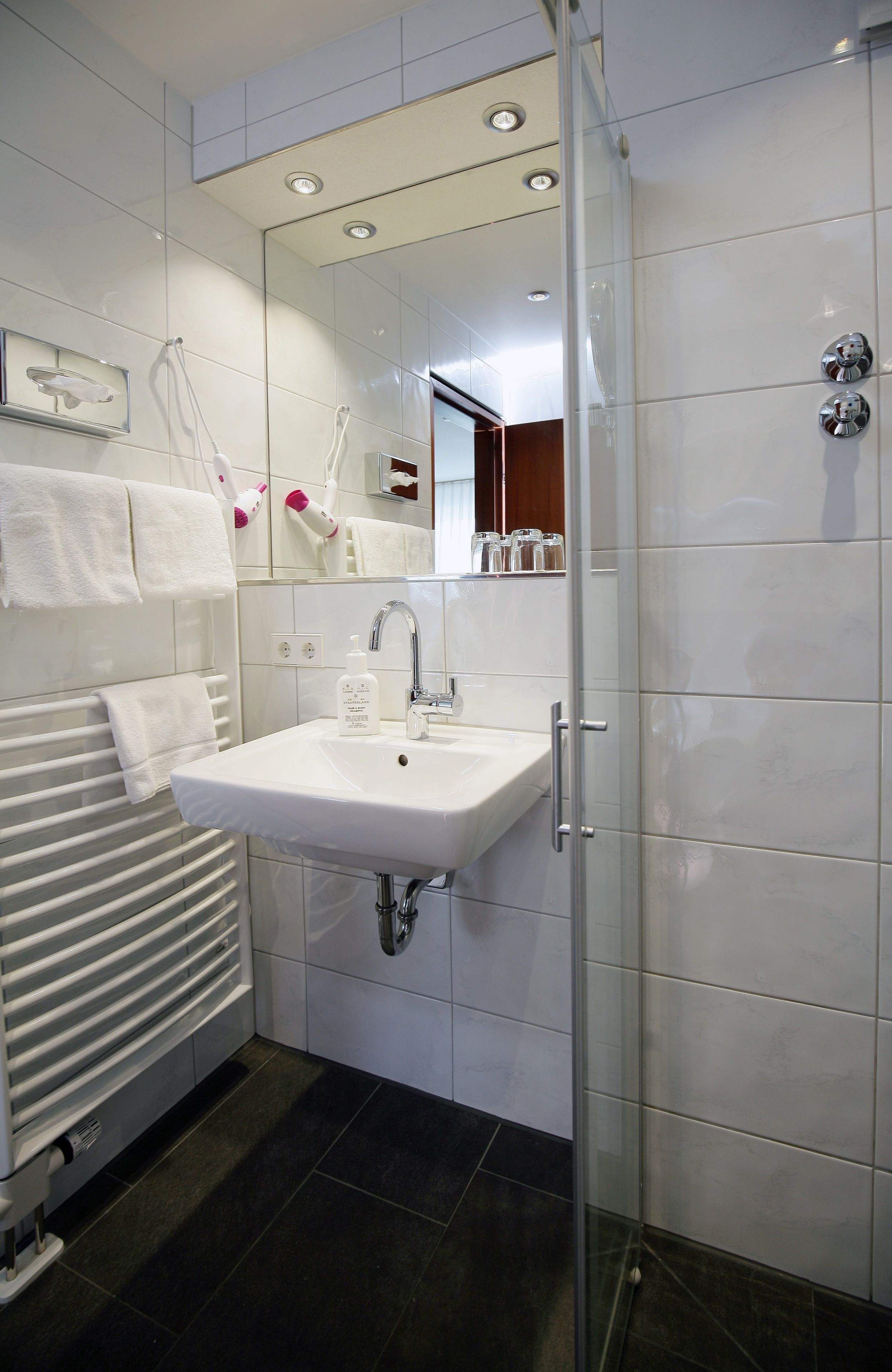 Un baño moderno con un lavamanos blanco, una cabina de ducha de vidrio y un perchero con toallas blancas.