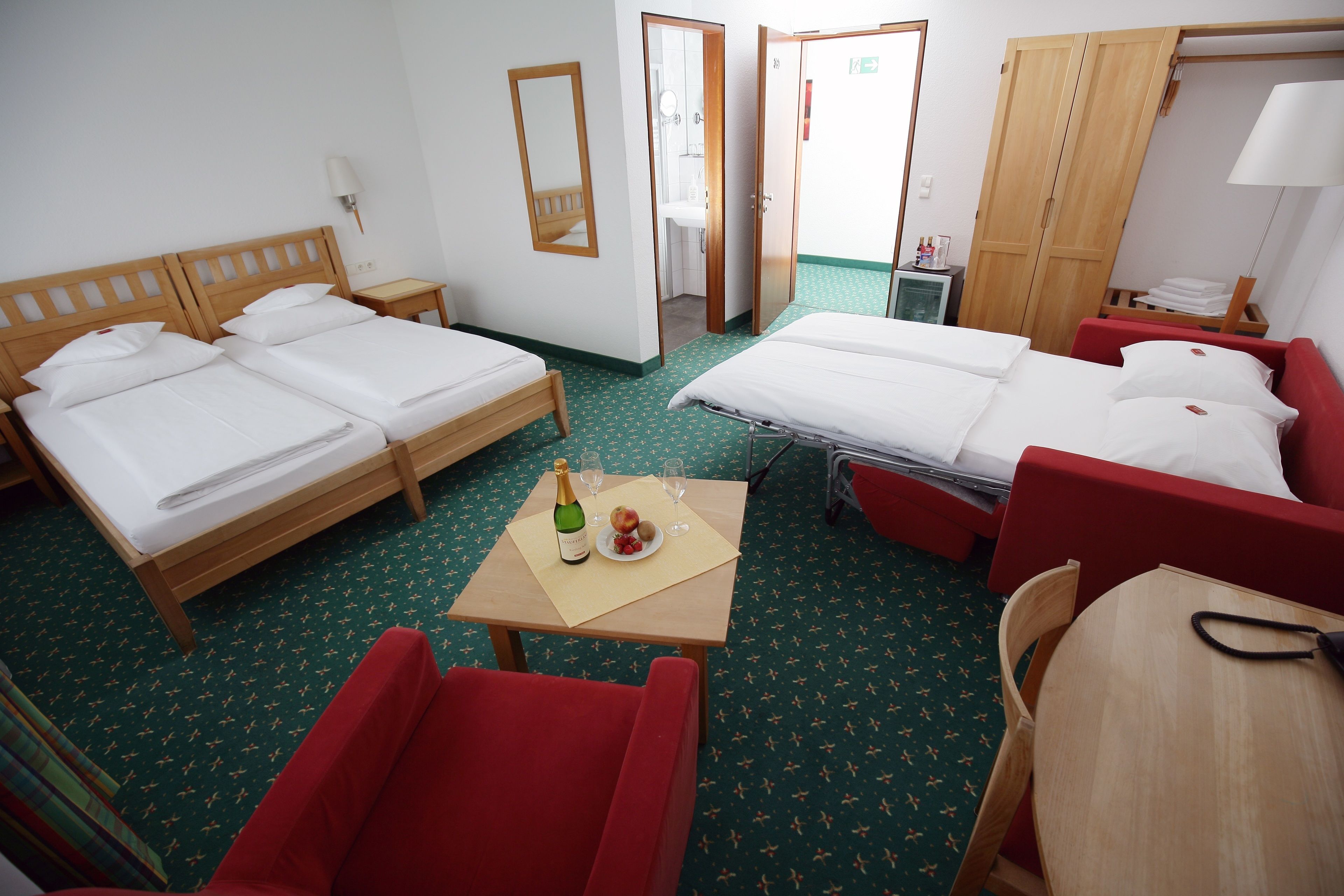 A hotel room with two single beds, a red sofa, a small table with a bottle and glasses, and a mirror on the wall.