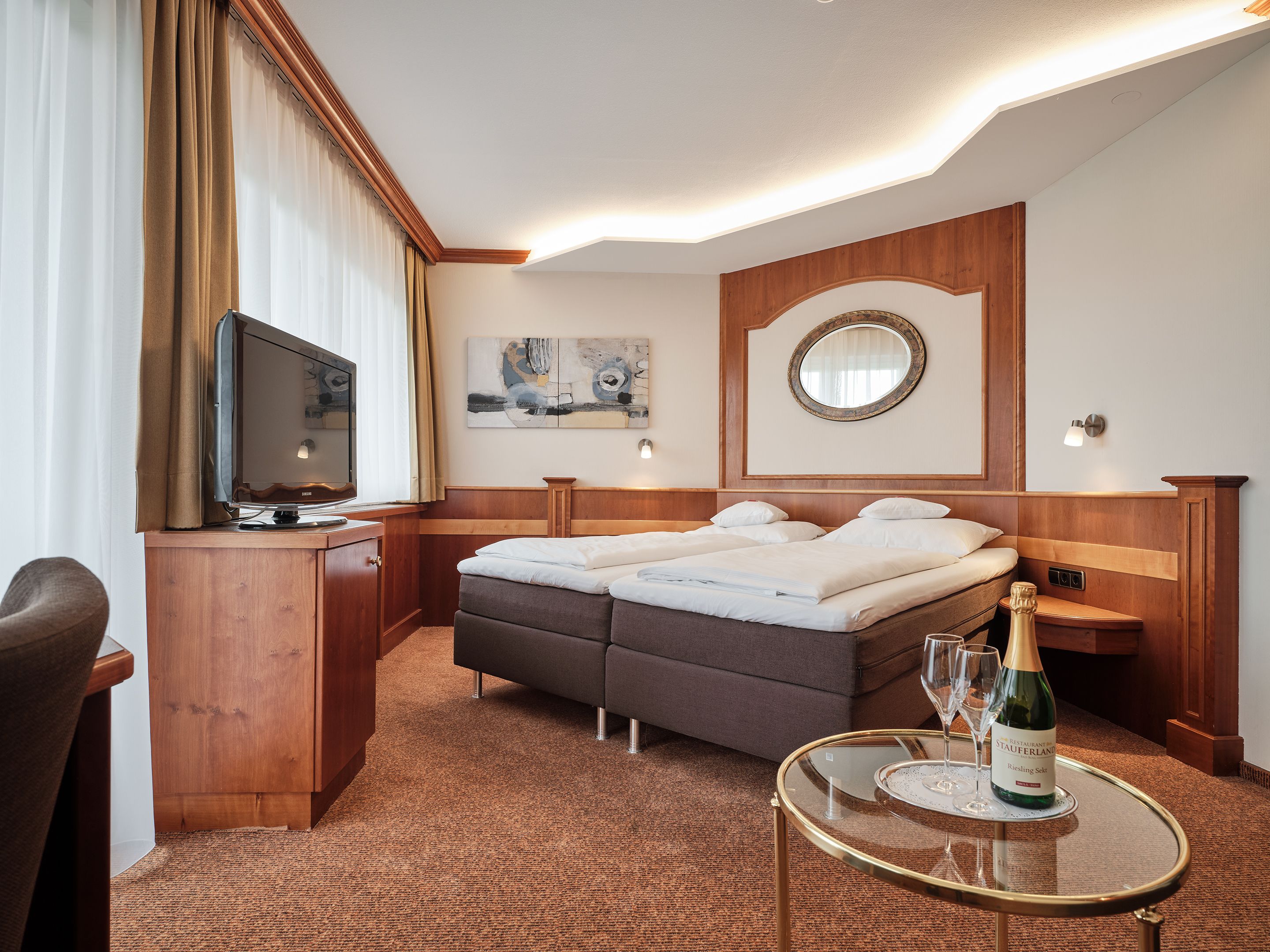 A hotel room with a bed, a desk with a television, a small table with a bottle of wine, and a mirror on the wall.
