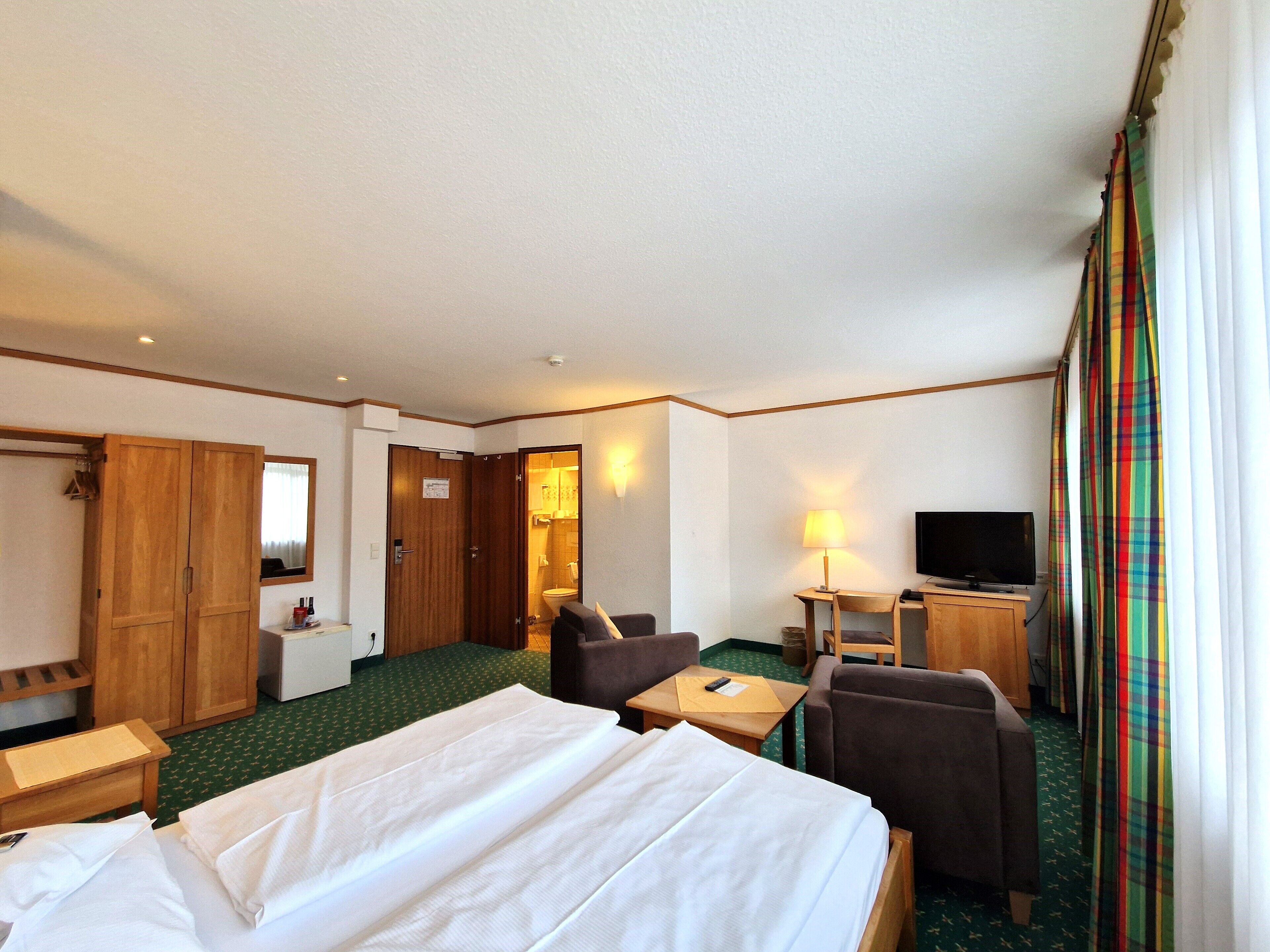 A hotel room with a bed, two chairs, a desk, a television, and a bathroom visible through an open door.