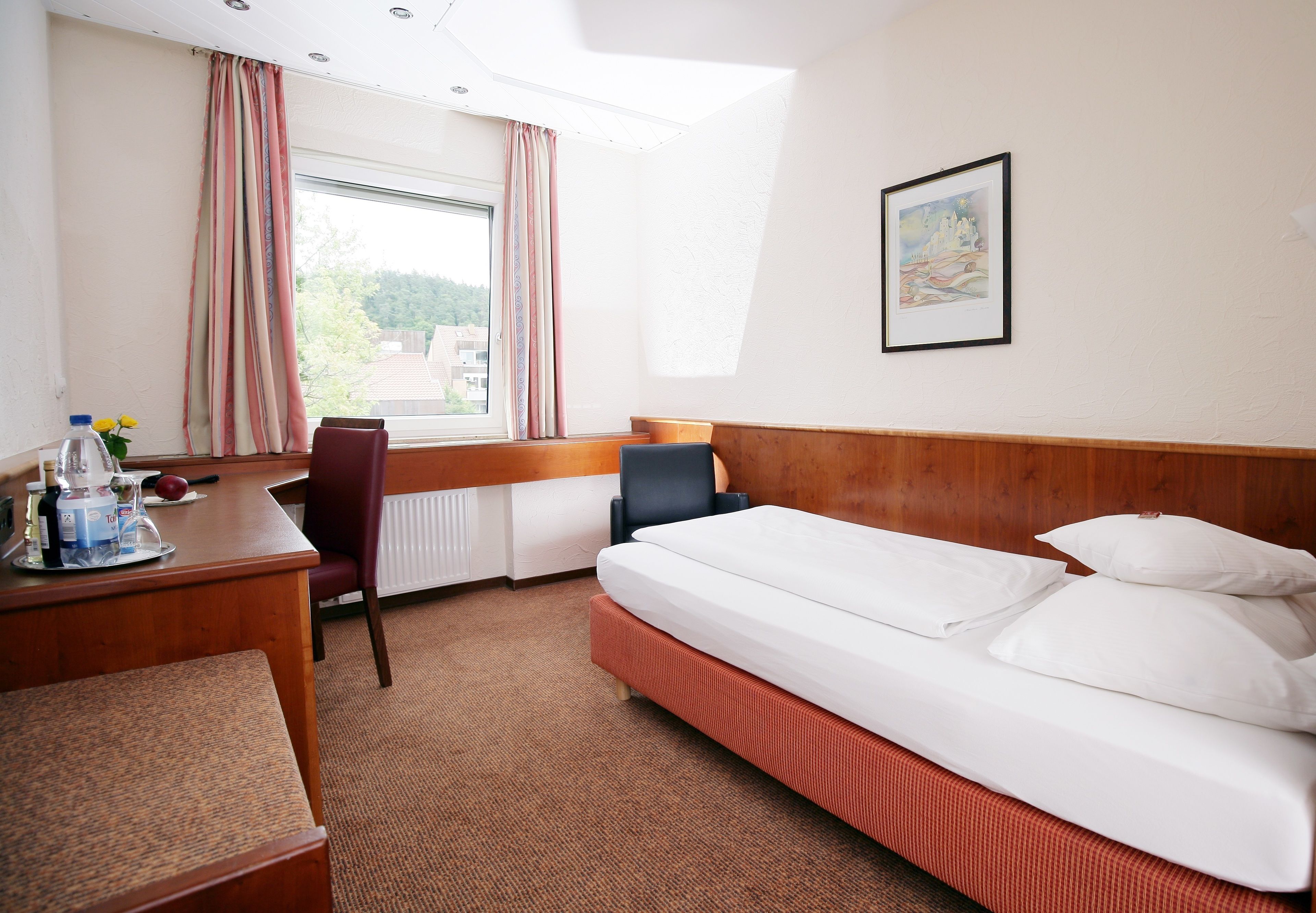 Ein Hotelzimmer mit Bett, Schreibtisch, Stuhl, einem Fenster mit Vorhängen und einem gerahmten Bild an der Wand.