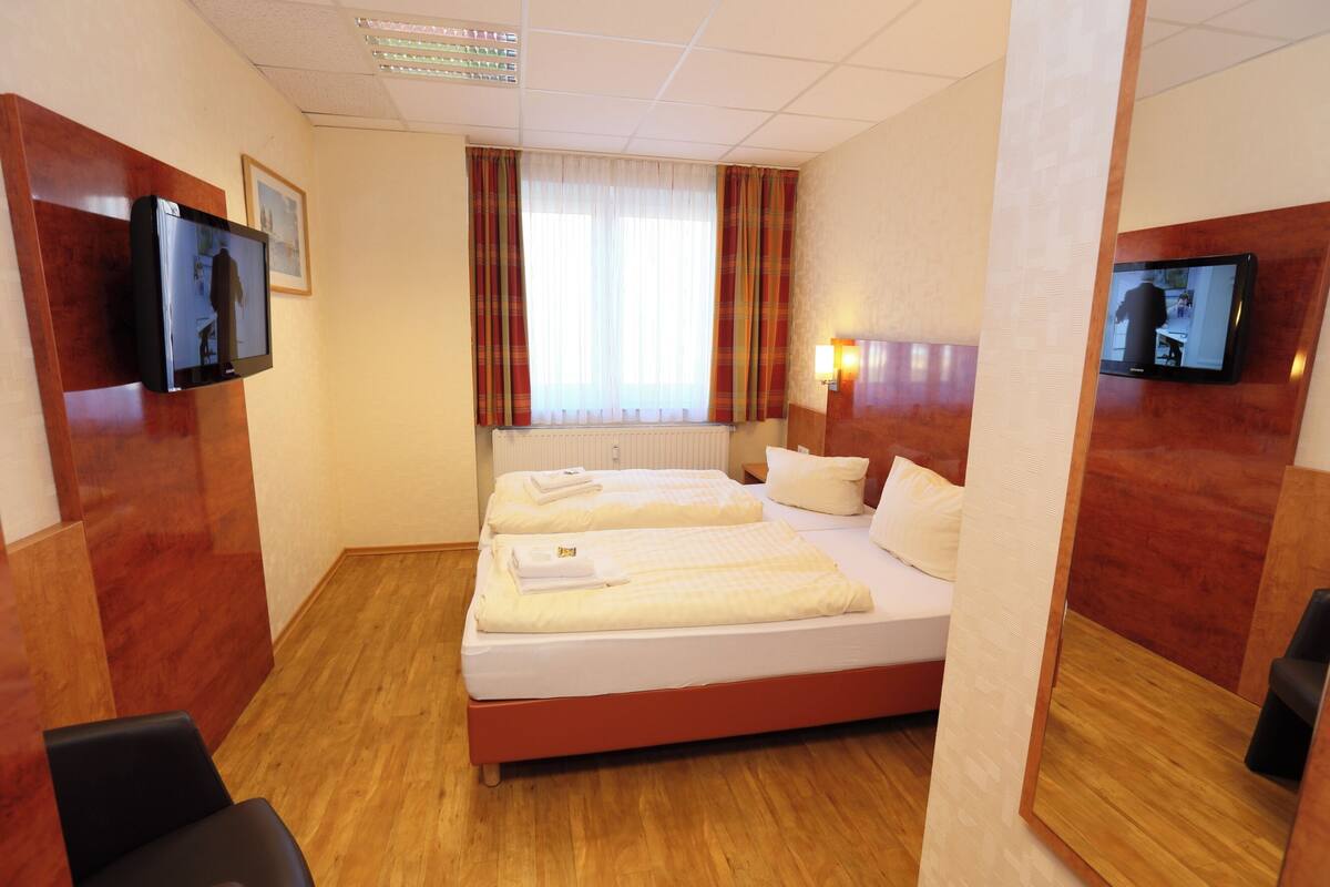 A hotel room with a large bed, two bedside tables, a TV mounted on the wall, and a window with curtains.