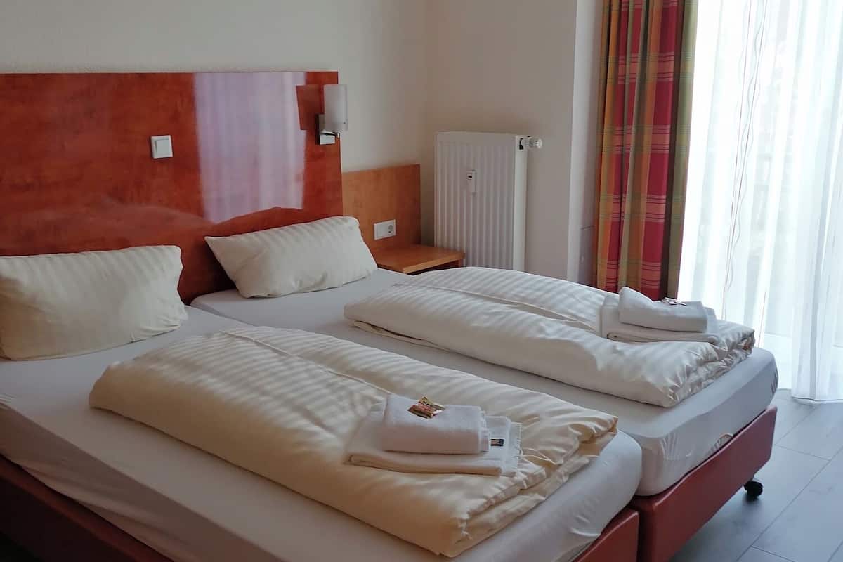 A hotel room with two beds, a wooden headboard, a radiator, and a window with curtains.