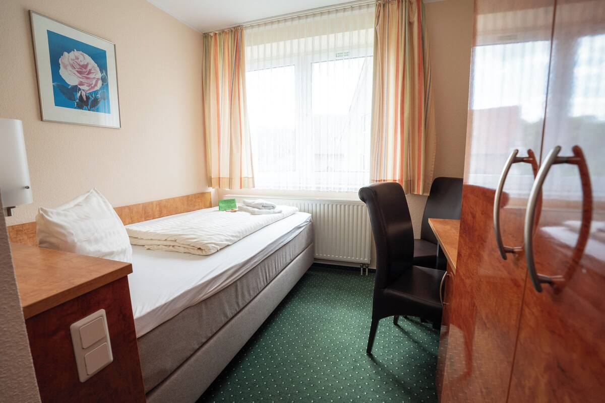 A hotel room with a bed, a desk, a chair, a radiator, and a window with curtains.