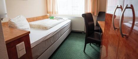 A hotel room with a bed, a desk, a chair, a radiator, and a window with curtains.