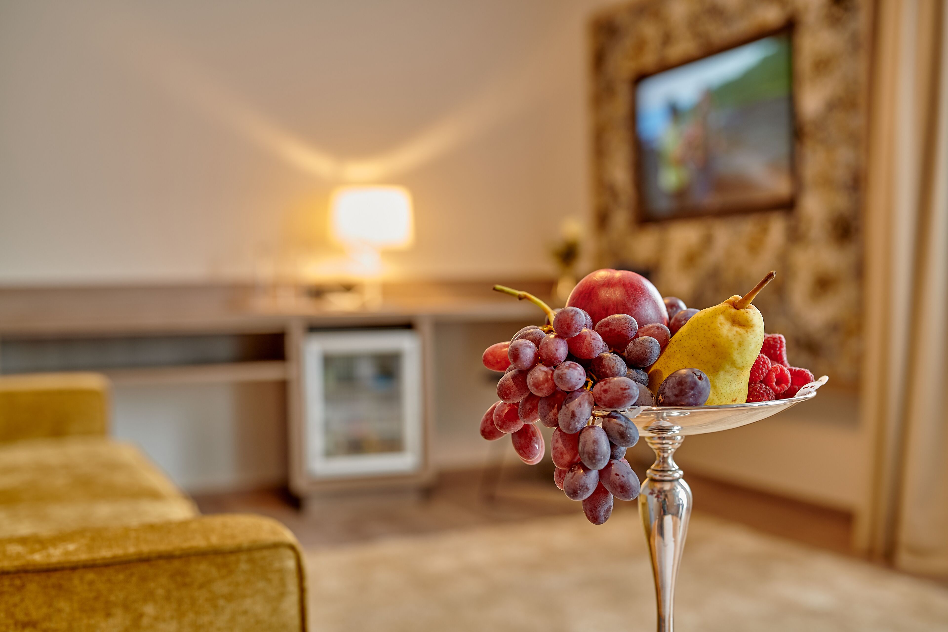 A fruit arrangement on a silver tray in a room with a lamp, a framed picture on the wall, and a yellow couch.