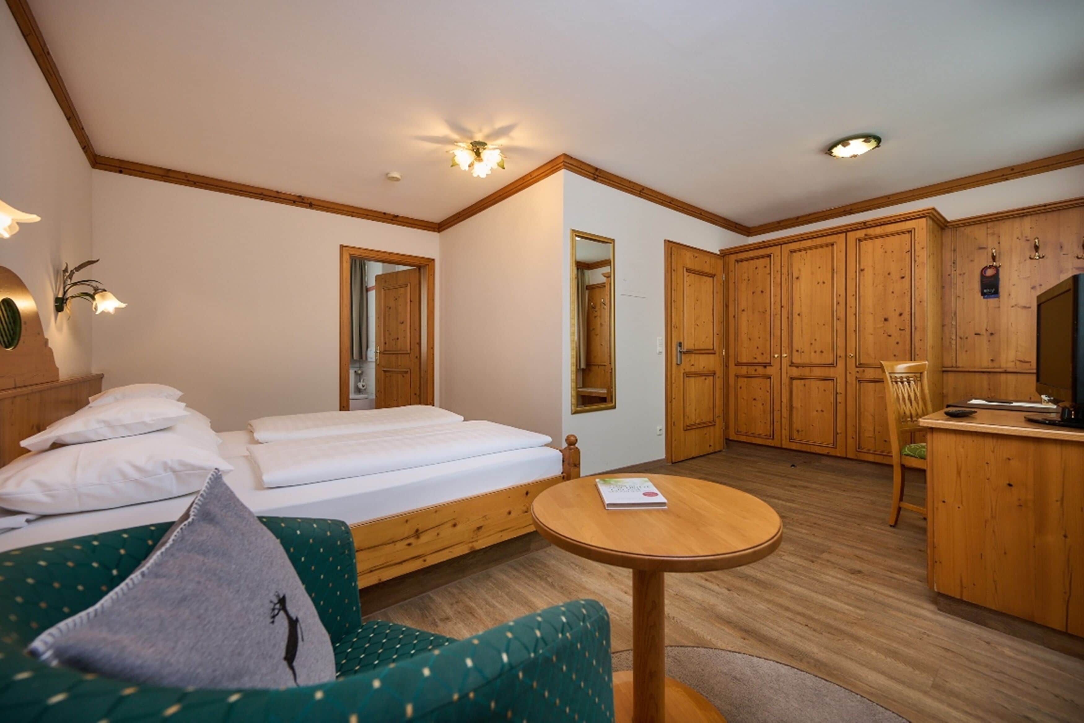 Habitación de hotel con cama, sofá verde, mesa redonda de madera, televisor, armarios de madera y un espejo.