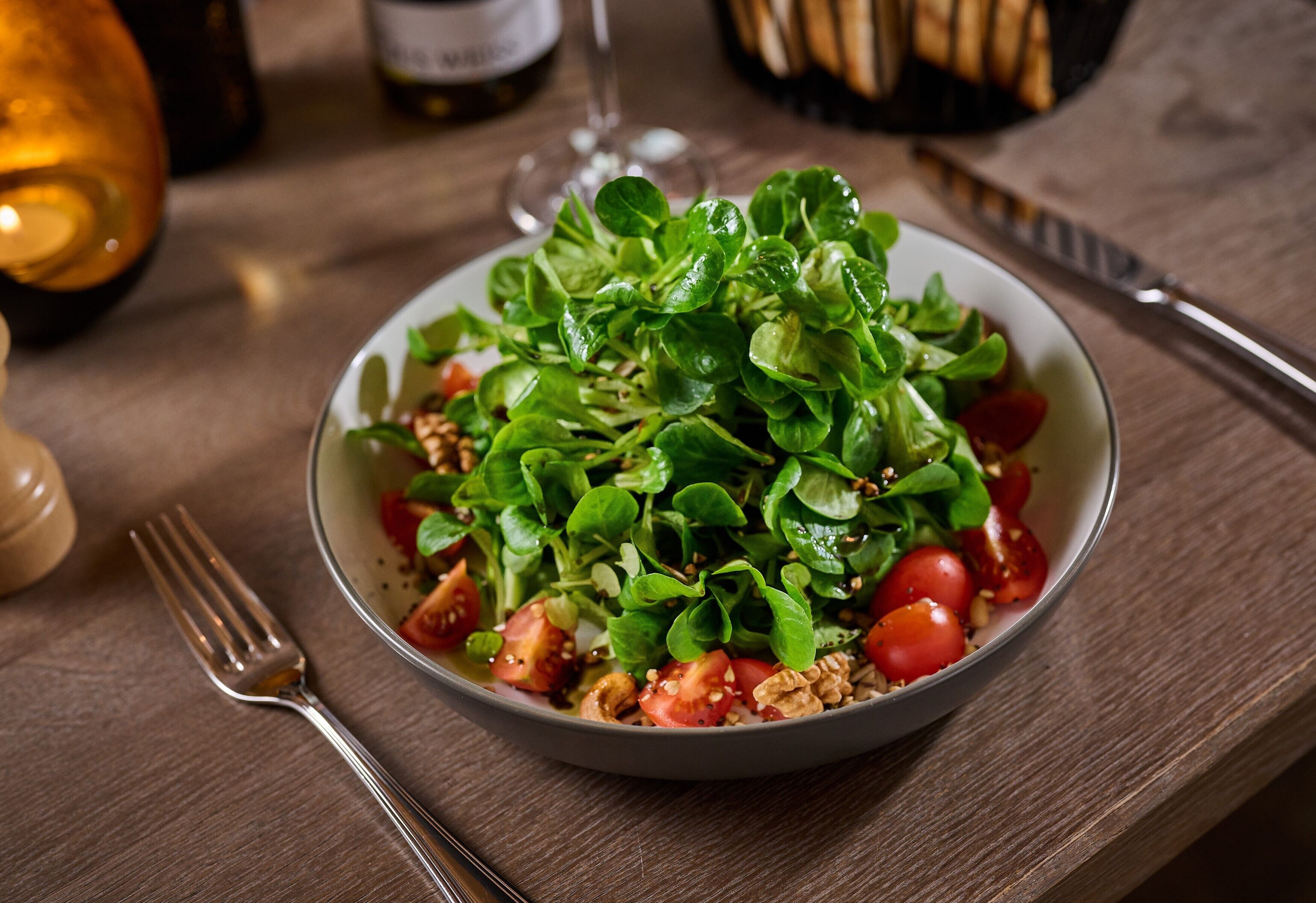 A bowl of salad with various greens and tomatoes on a wooden table.