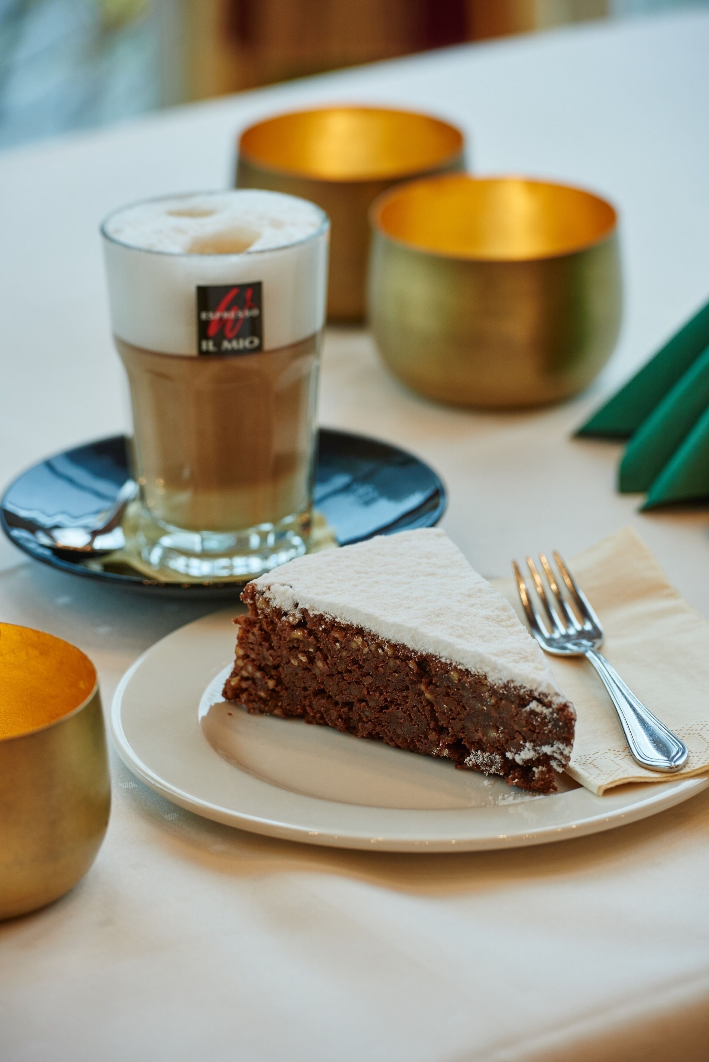 A slice of cake on a plate with a fork, a glass of coffee, and two golden bowls on a table.