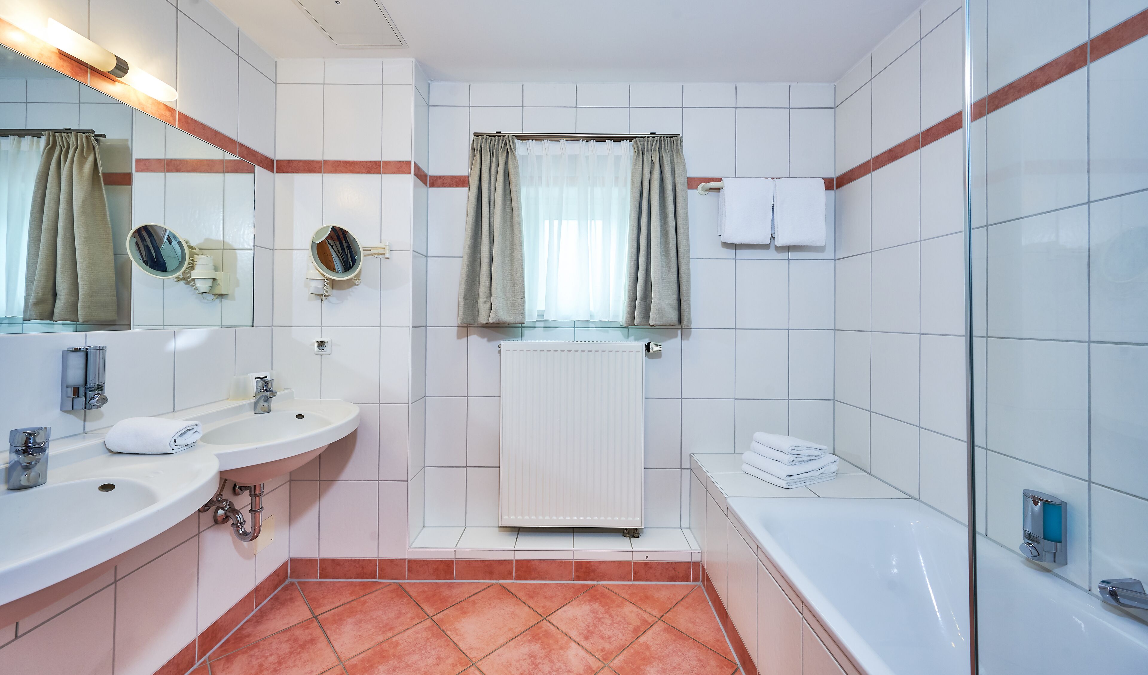 A bathroom with white tiled walls, a white sink, a mirror, a window with curtains, a radiator, and a bathtub.
