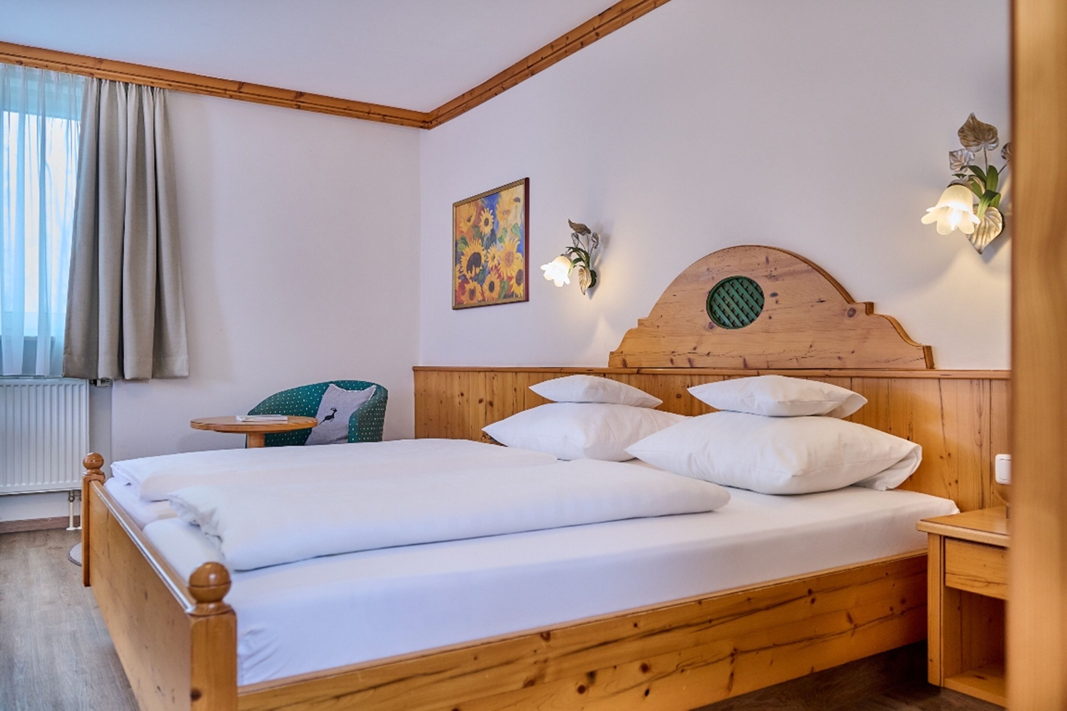 A wooden bed with white bedding, a small bedside table, a wall-mounted light fixture, and a painting of sunflowers.