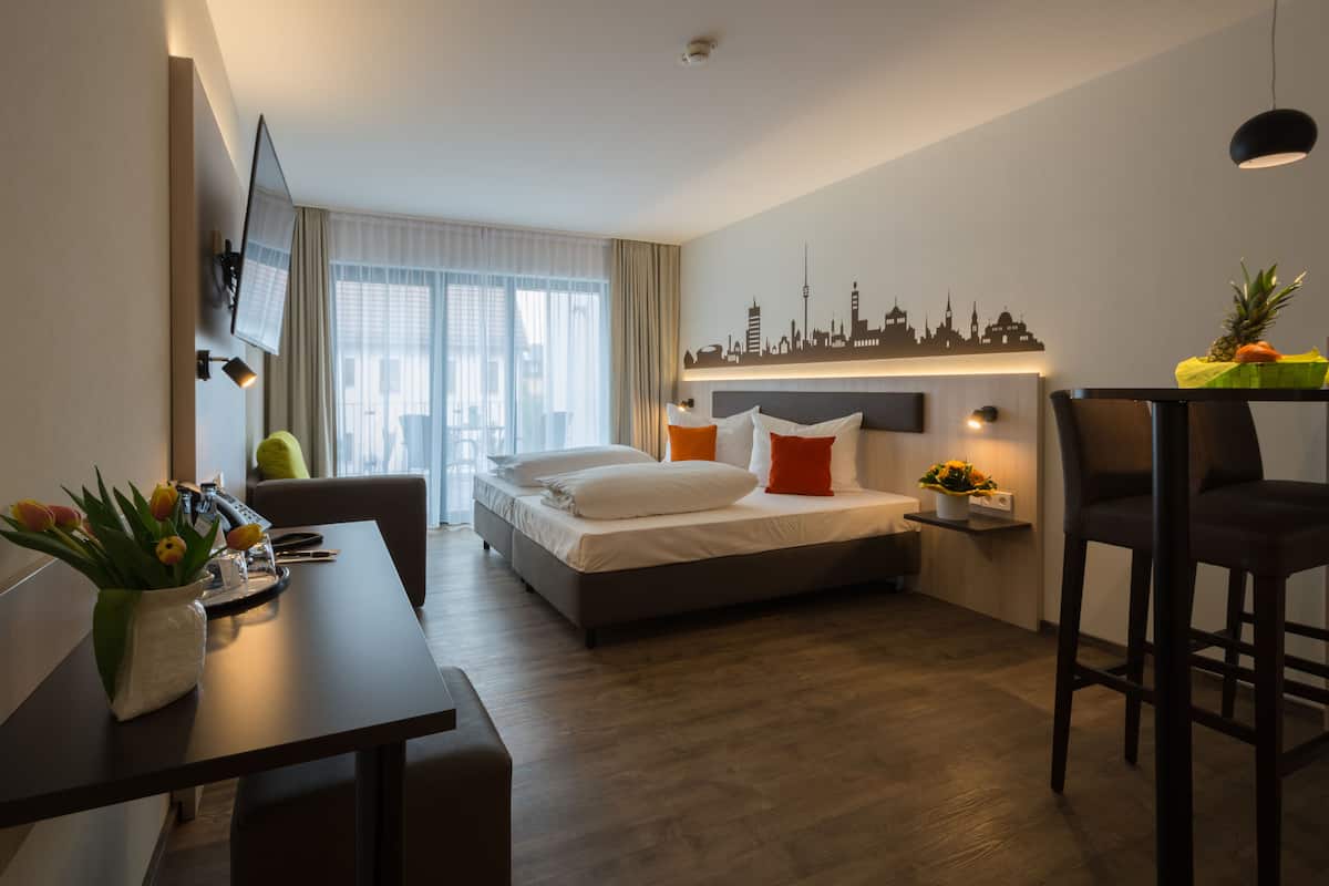 Habitación de hotel moderna con cama, escritorio, silla, televisor y un mural con vista a un horizonte urbano.