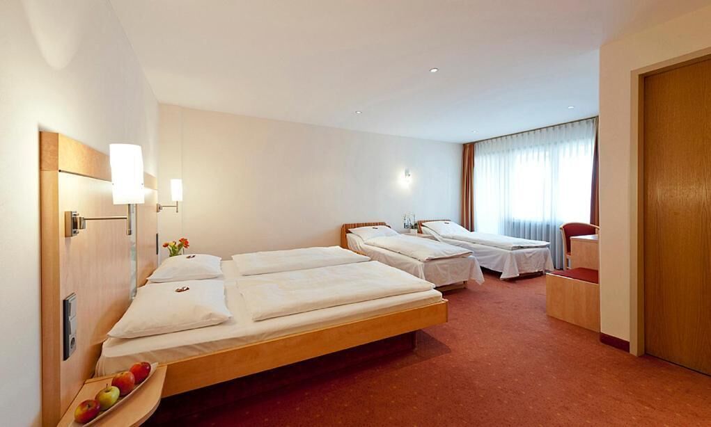 A hotel room with two single beds, a wooden desk, a chair, a small table with apples, and a window with curtains.