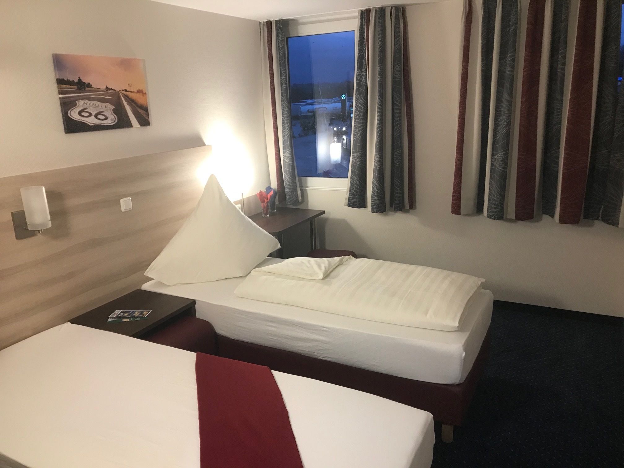 A hotel room with a bed, a nightstand, a window with curtains, and a wall-mounted picture of Route 66.