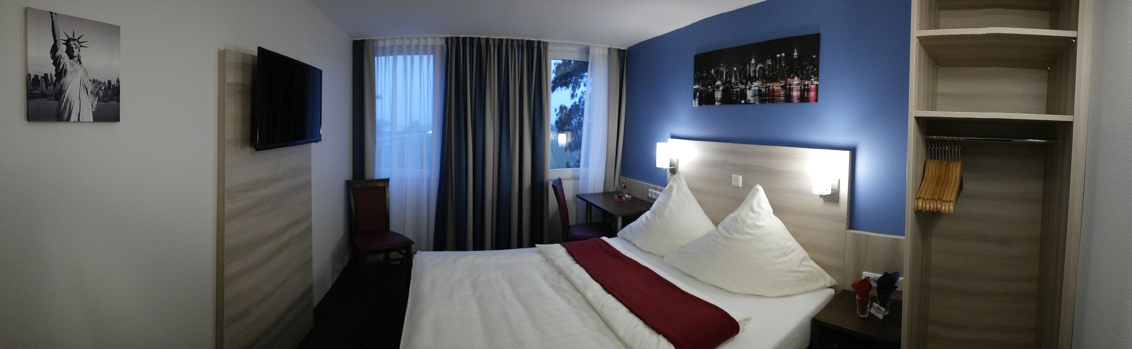 A hotel room with a bed, a TV, a desk, a chair, a wardrobe, and a window with curtains.