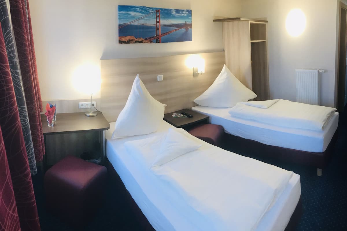 A hotel room with two beds, a desk, a chair, a wall-mounted TV, and a painting of the Golden Gate Bridge.