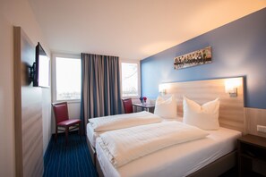 Una habitación de hotel con una cama grande, un escritorio con silla, un televisor montado en la pared y una ventana con cortinas.