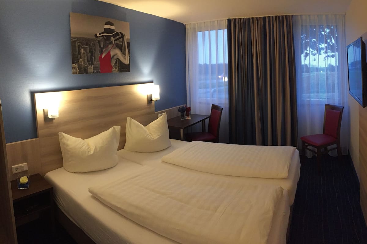 A hotel room with a large bed, a desk with a chair, a flat-screen TV mounted on the wall, and a window with curtains.