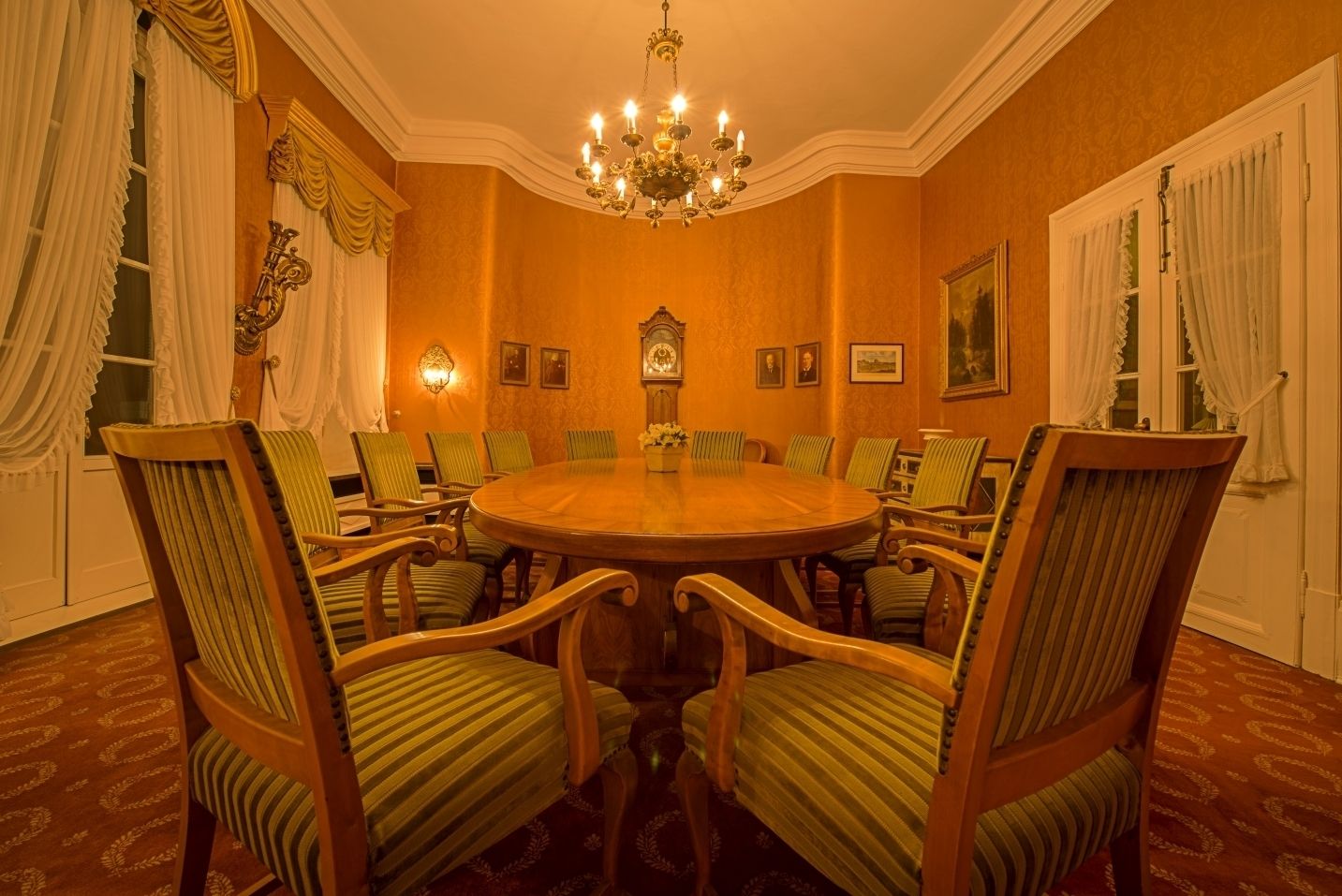 Una sala riunioni formale con un tavolo rotondo, sedie imbottite in verde, orologi a parete e un lampadario.