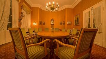 Een zakelijke vergaderruimte met een ronde tafel, groene bekleden stoelen, wandklokken en een kroonluchter.