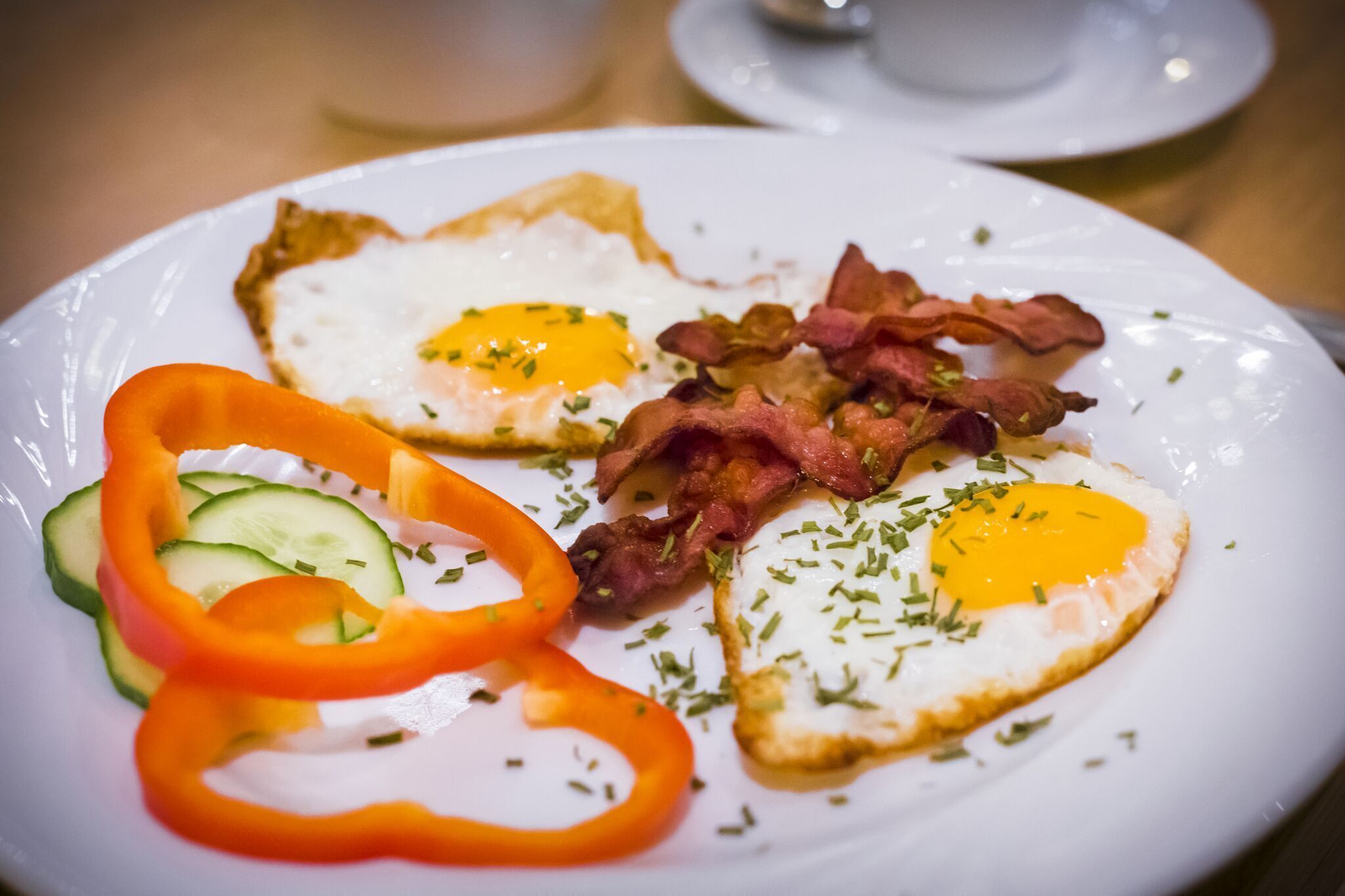 Une assiette contenant deux œufs au plat, des tranches de bacon, des rondelles de concombre et des lanières de poivron rouge.