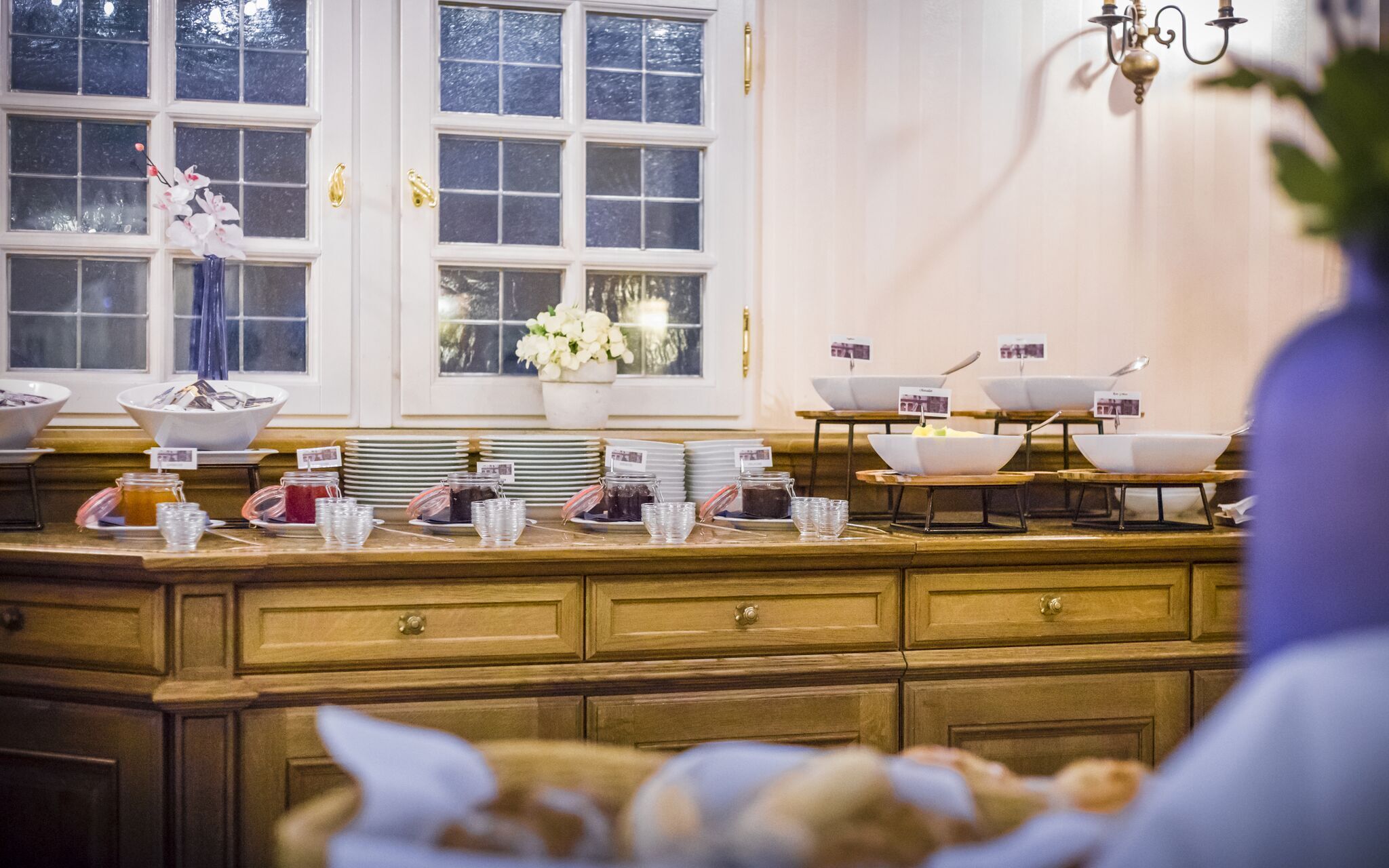 A buffet setup with multiple bowls and plates, a window with flowers, and a chandelier.