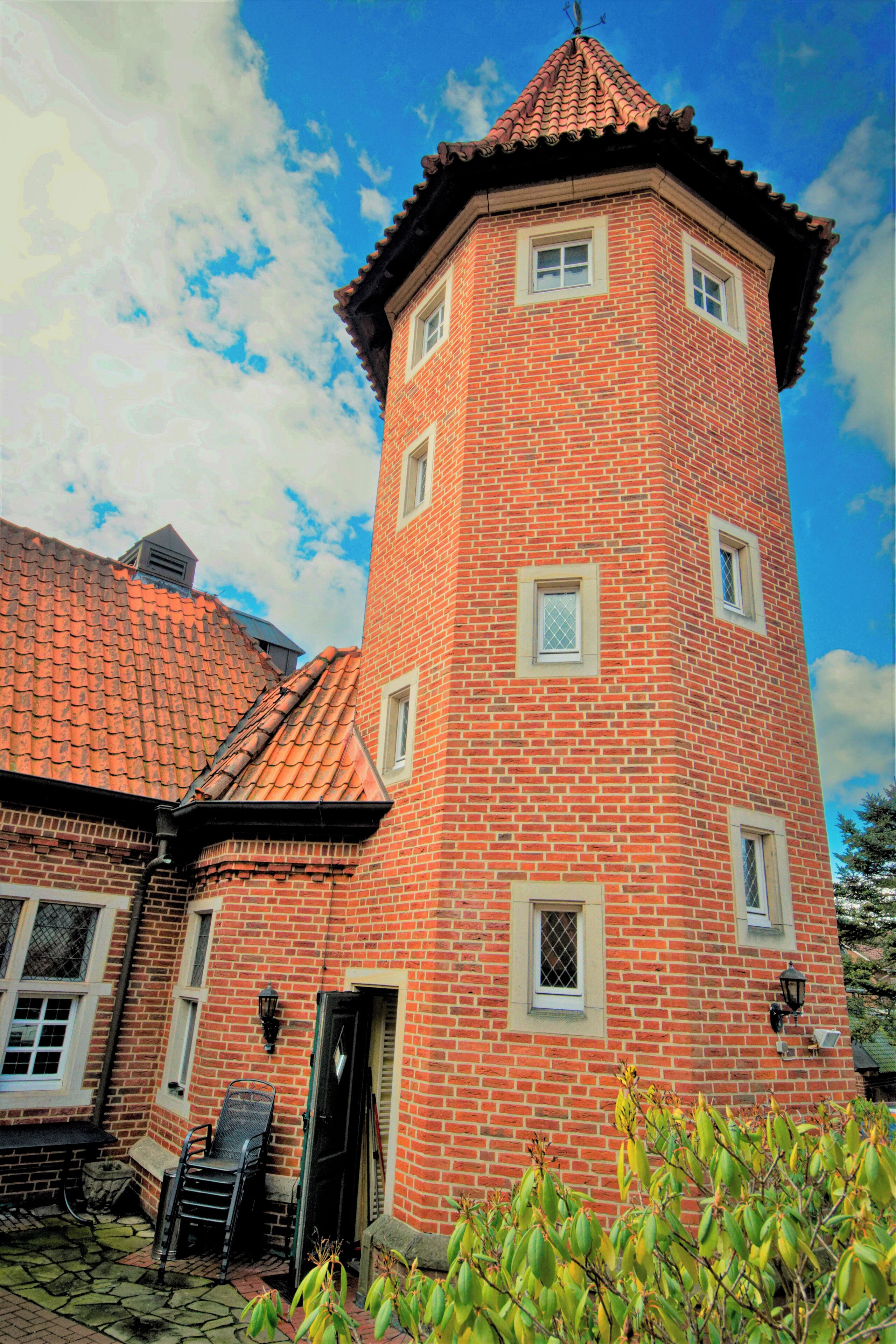 Una torre de ladrillo rojo con múltiples ventanas y una puerta, rodeada por un jardín con plantas de banano.