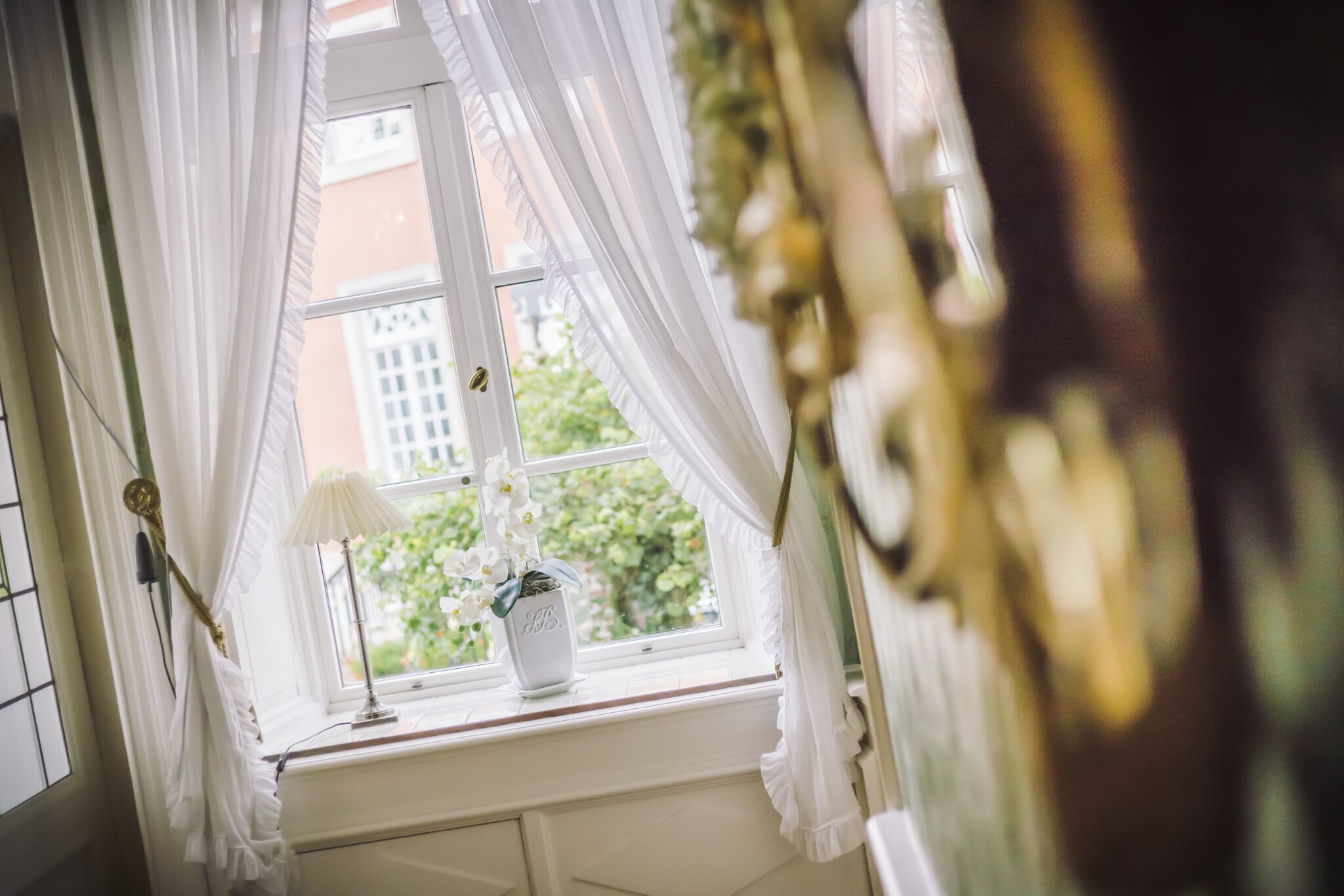 A window with white curtains, a lamp, and a vase with flowers.