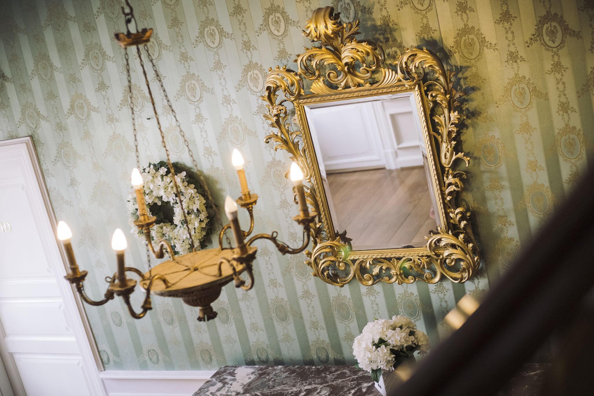 En kristallkrona med flera ljuskällor som liknar ljus, en spegel med en utsmyckad guldfram och en blomsterdekoration på väggen.