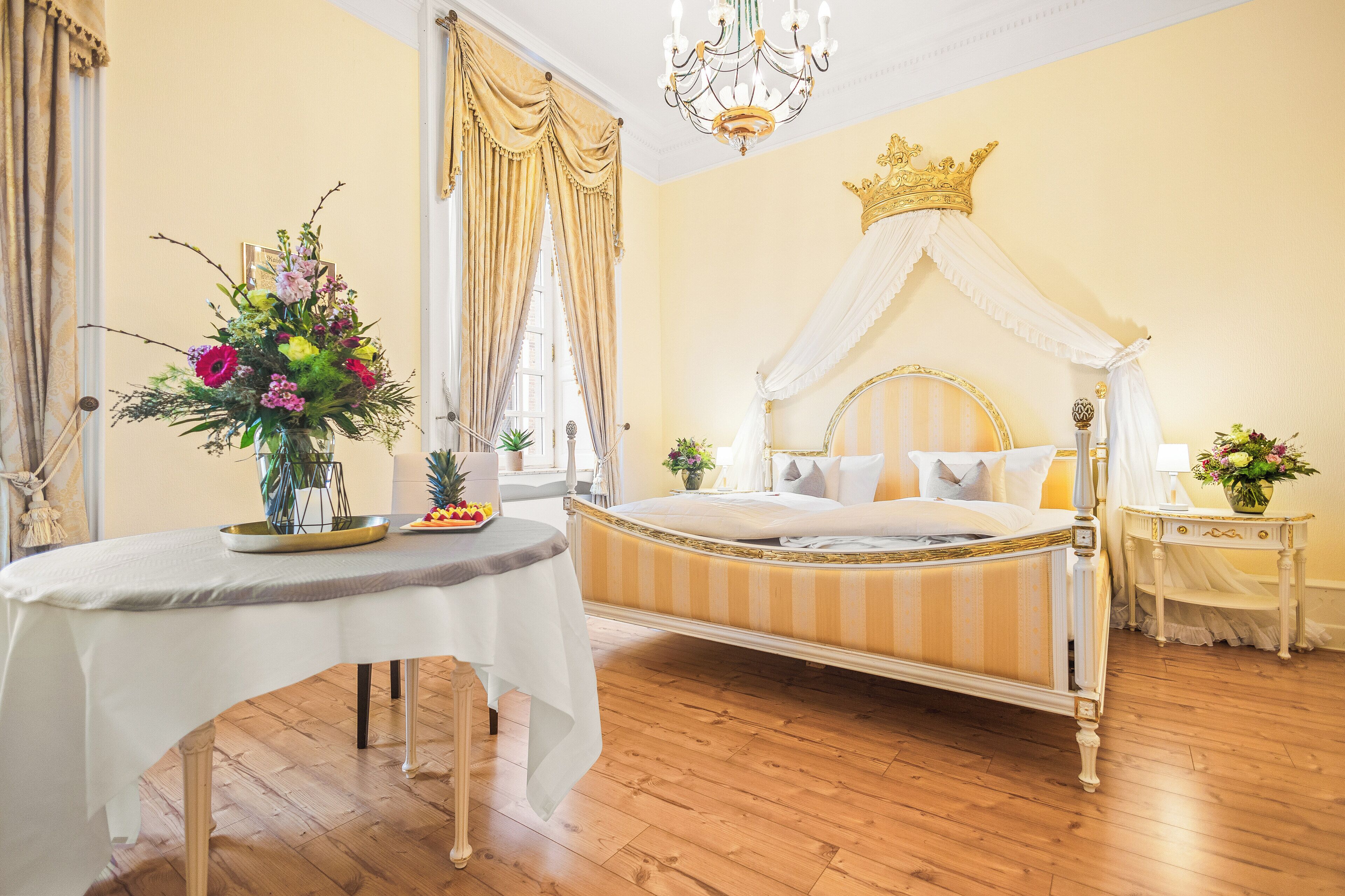 A bedroom with a four-poster bed, a chandelier, and a floral arrangement on the bedside table.