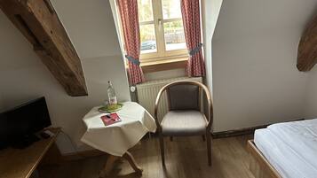 Ein kleiner Raum mit einem Bett, einem Holztisch, einem Stuhl, einem Tisch mit Flasche und Glas sowie einem Fenster mit Vorhängen.