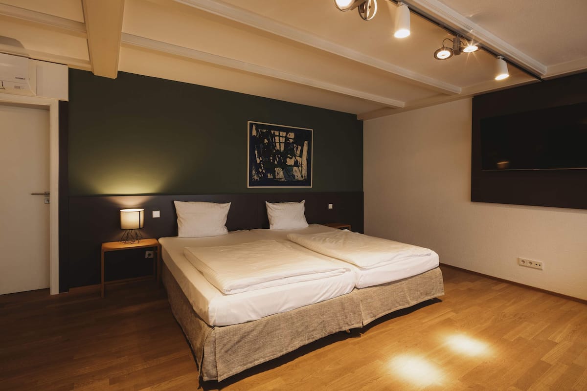 A bedroom with a double bed, a bedside table with a lamp, a wall-mounted TV, and a ceiling light fixture.