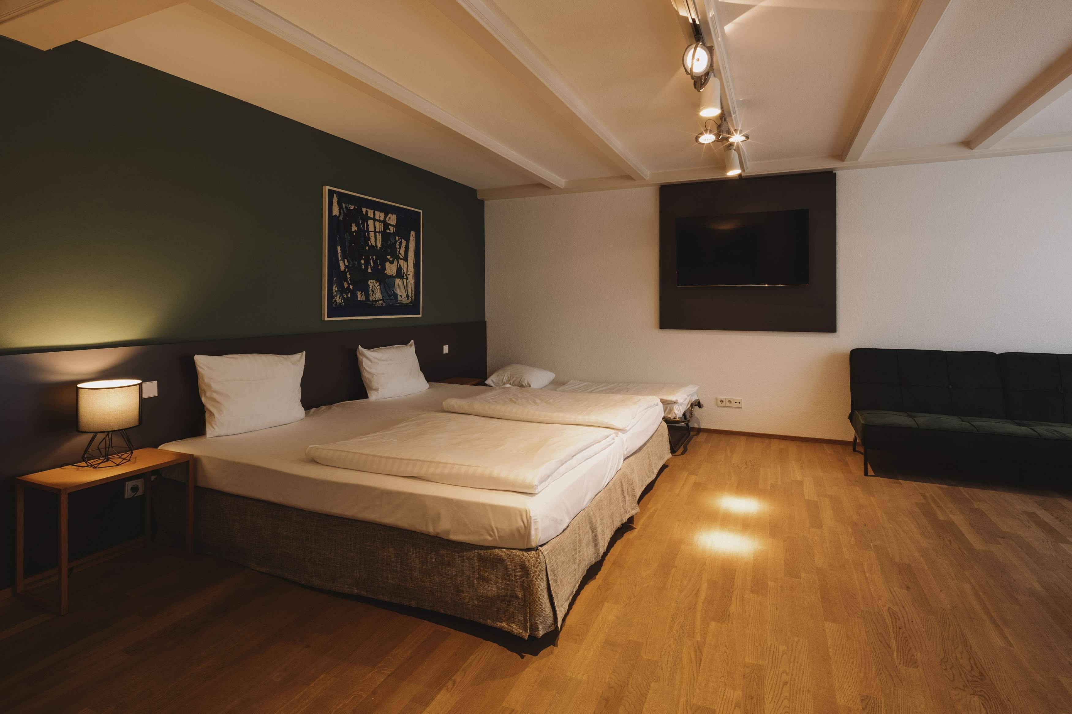 Une chambre d’hôtel avec un grand lit, une table de chevet avec une lampe, un mur d’accent de couleur vert foncé, un téléviseur fixé au mur et un canapé vert.