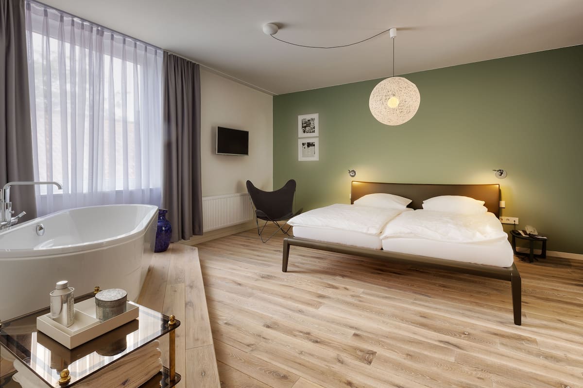 A modern bedroom with a large bathtub, a bed with white linens, a wooden floor, and a green accent wall.