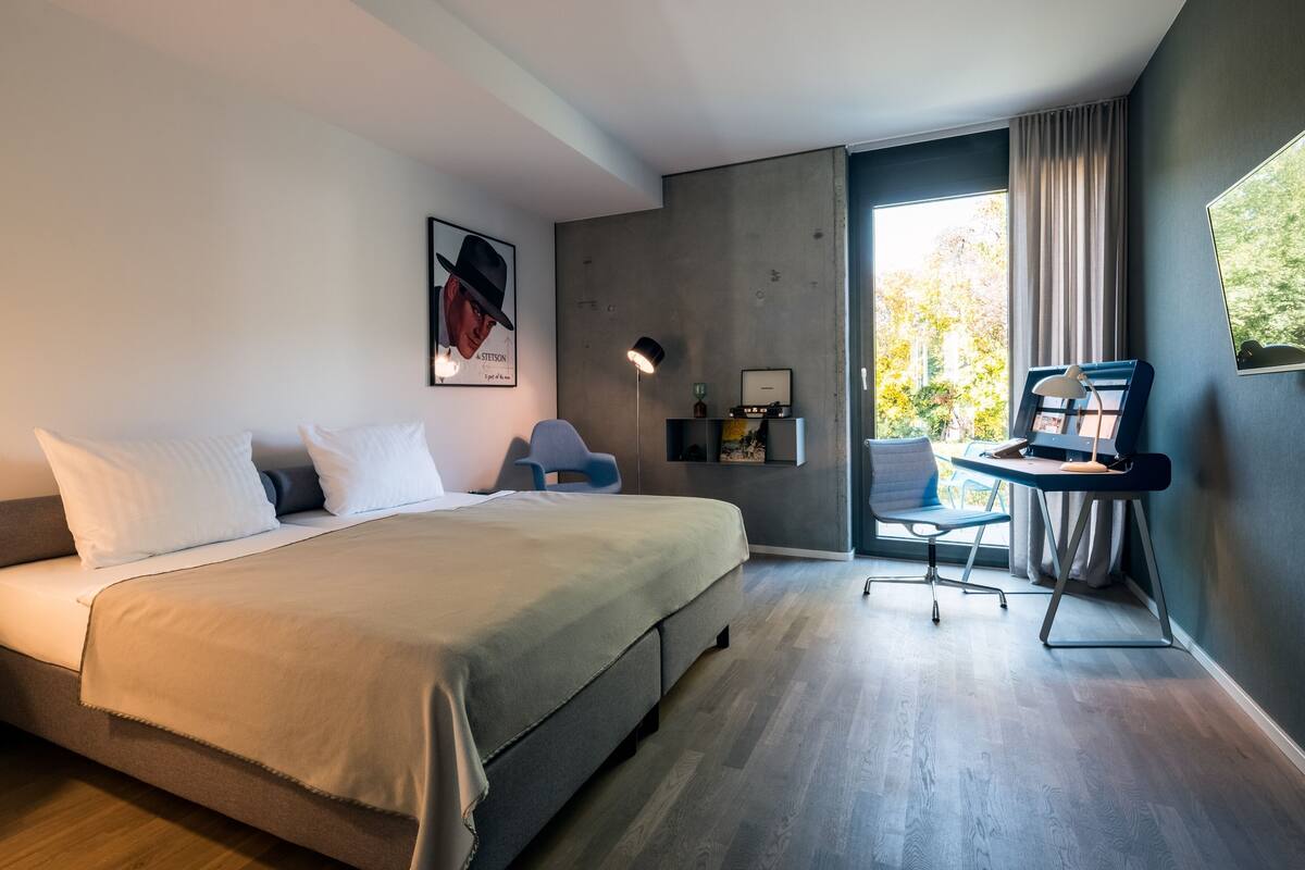 A modern bedroom with a bed, a desk, and a chair. There is a window with curtains, a picture on the wall, and a lamp.