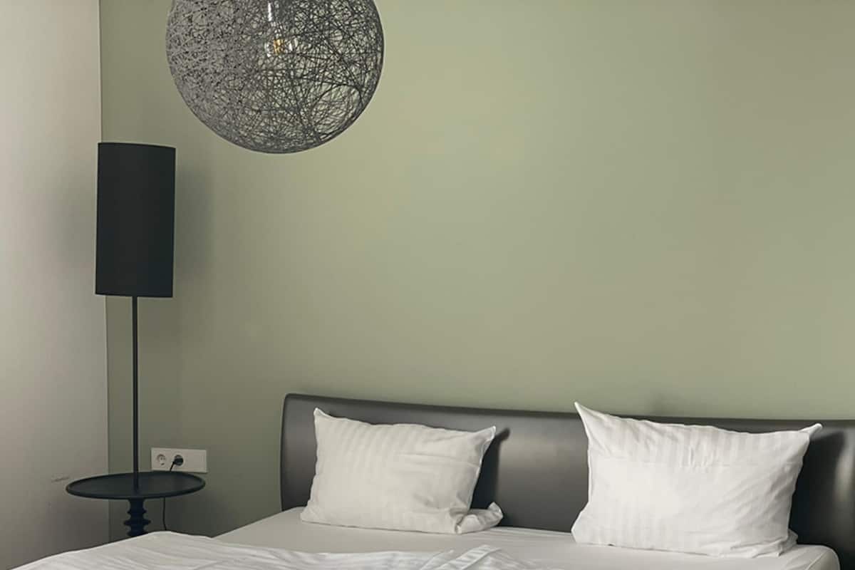 A modern hotel room with a bed, two pillows, a round hanging light fixture, and a small table with a lamp.