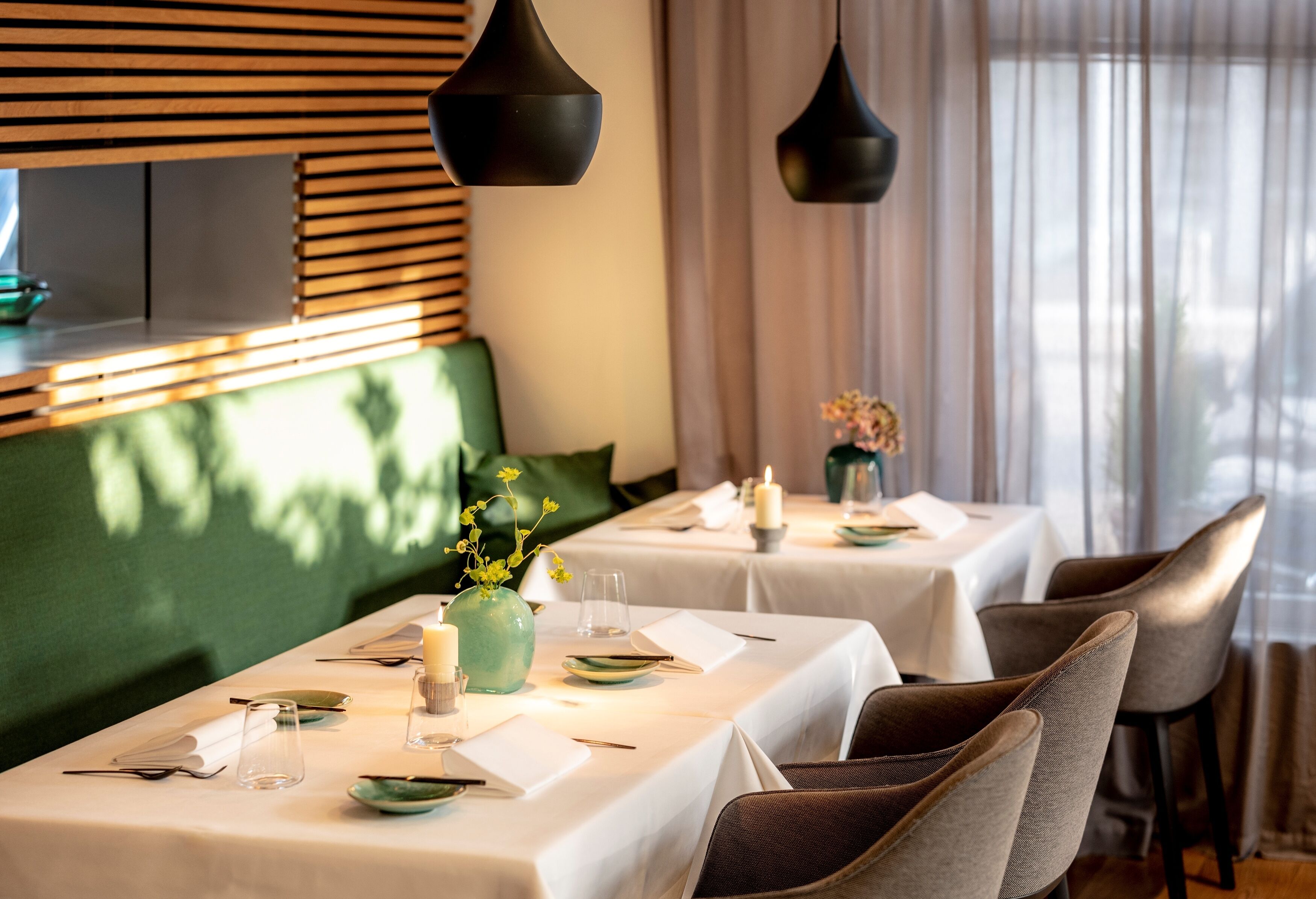 Un restaurant où les tables sont dressées pour le service, avec un coin salon vert capitonné, des nappes blanches et des luminaires suspendus noirs.