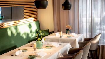 Un restaurant où les tables sont dressées pour le service, avec un coin salon vert capitonné, des nappes blanches et des luminaires suspendus noirs.