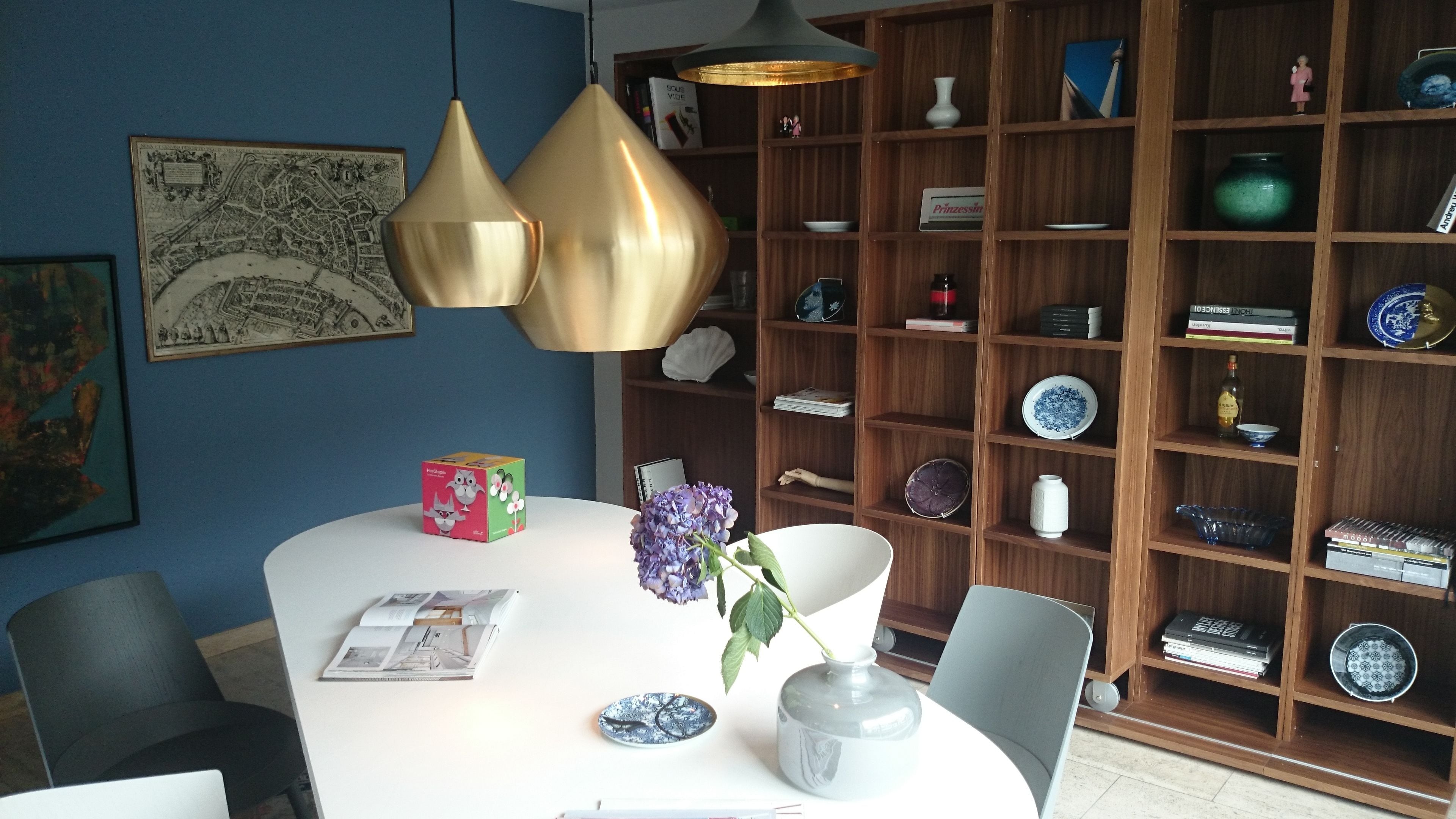 A modern dining area with a white table, a vase with purple flowers, and a bookshelf filled with books and decorative items.
