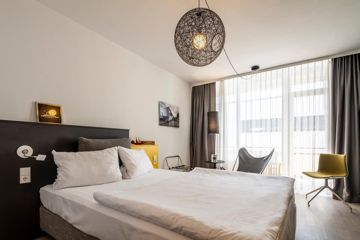 A modern hotel room with a large bed, a desk, a chair, and a large window with curtains.